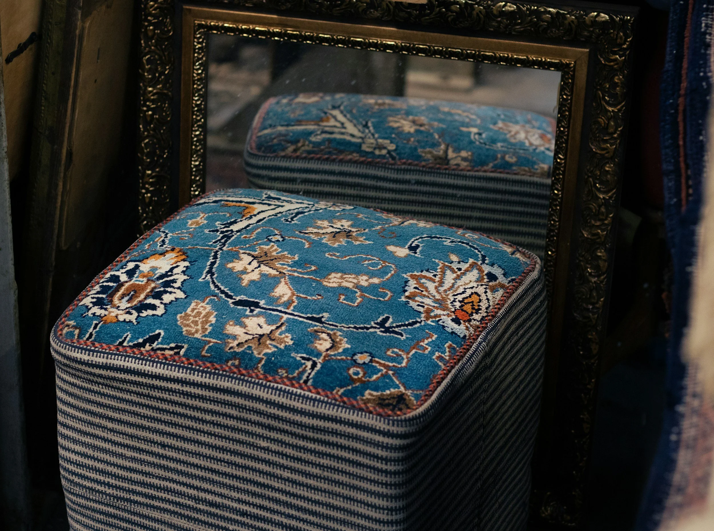 A stool with a blue, orange, and beige floral pattern on top, positioned in front of a decorative mirror with a dark carved frame.