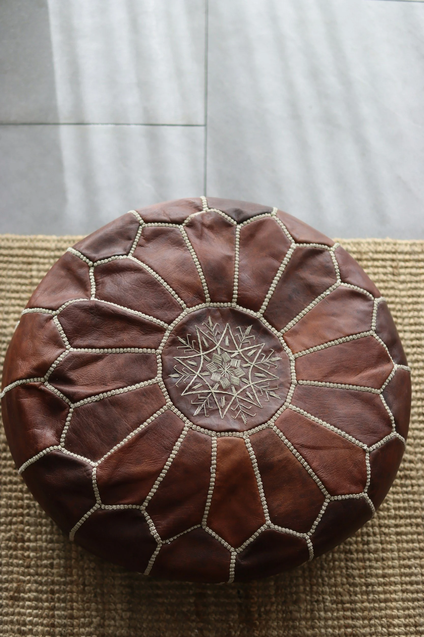 A round leather pouf with a floral and geometric embroidered design on top, placed on a woven mat against a plain wall.