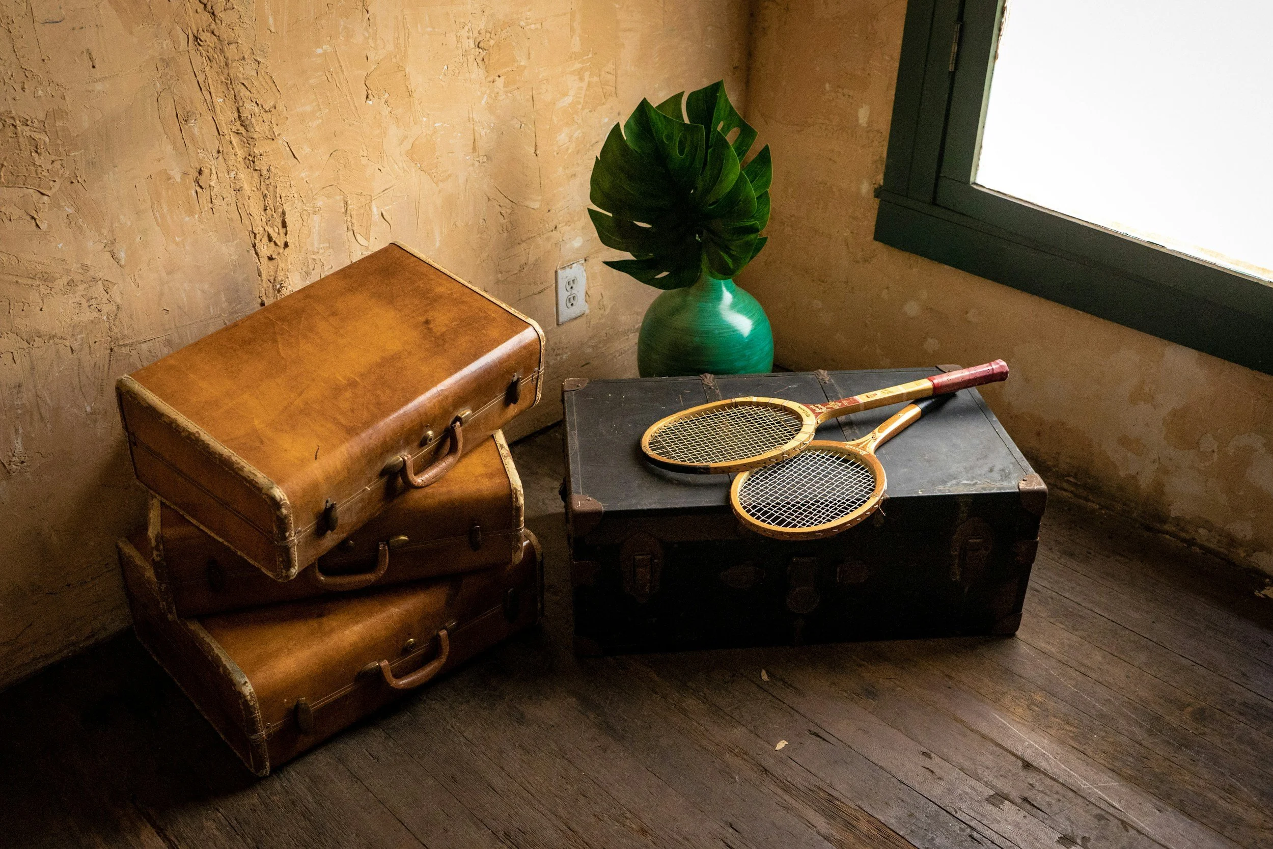 A vintage brown leather suitcase, a black wooden box, and two tennis rackets with orange frames and black strings arranged on a dark wooden floor near a textured beige wall and next to a window with black trim, with a green vase and large monstera leaf inside.