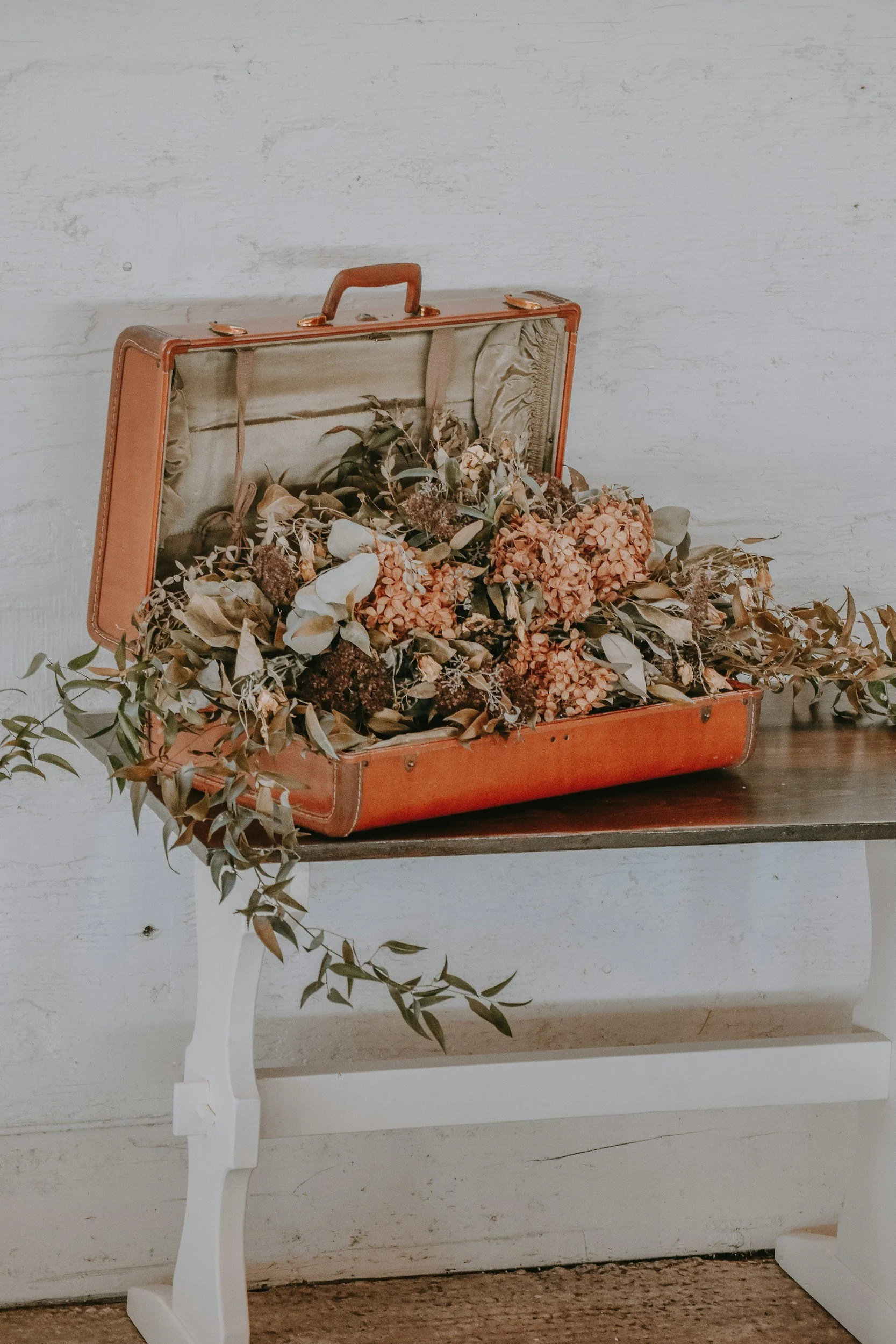 A vintage orange suitcase filled with dried flowers and foliage, open and resting on a wooden table. The background features a white textured wall.