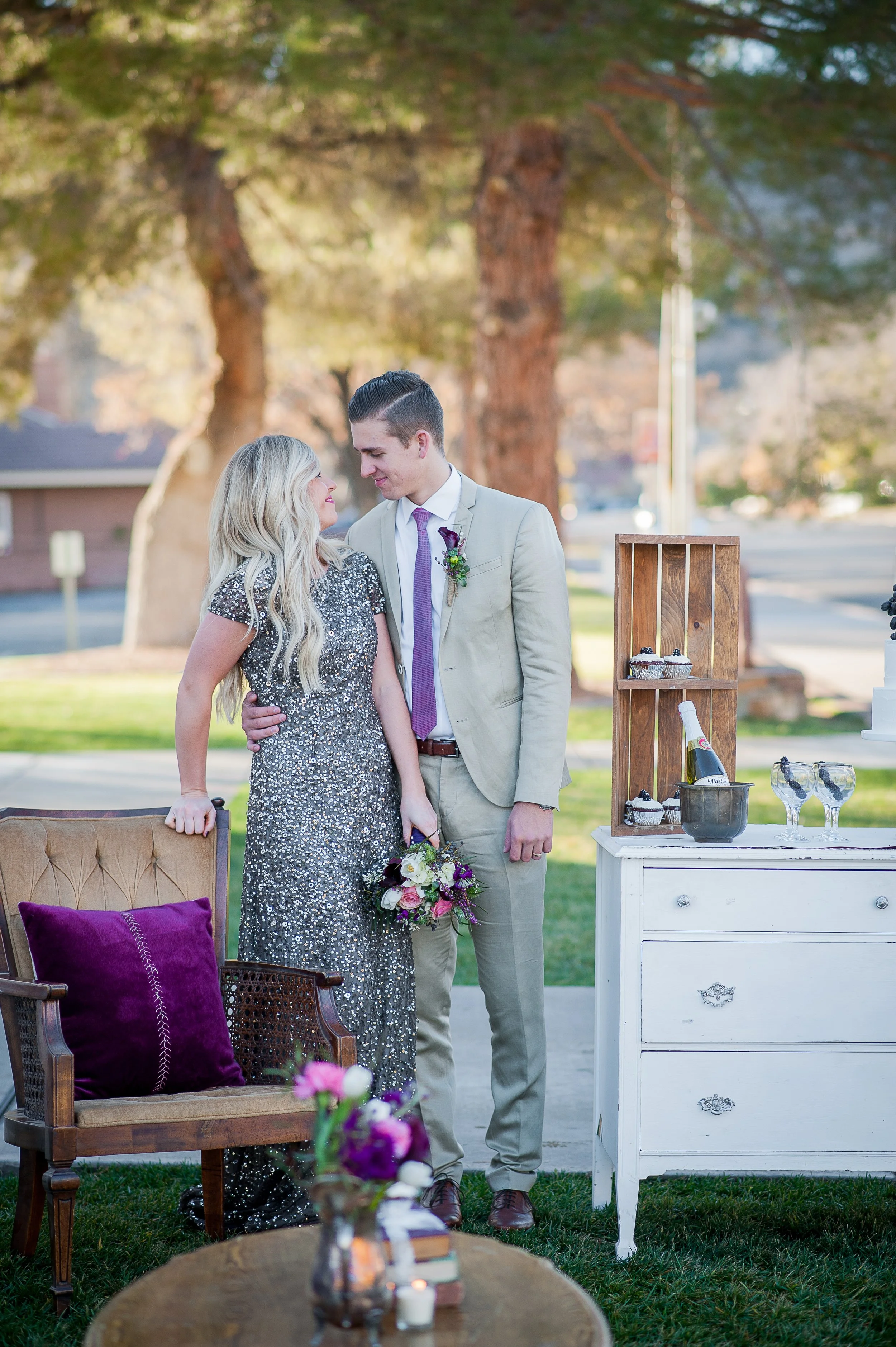 A couple dressed in formal attire standing close together outdoors under a tree, with flowers and cupcakes displayed nearby, suggesting a celebration or special occasion.