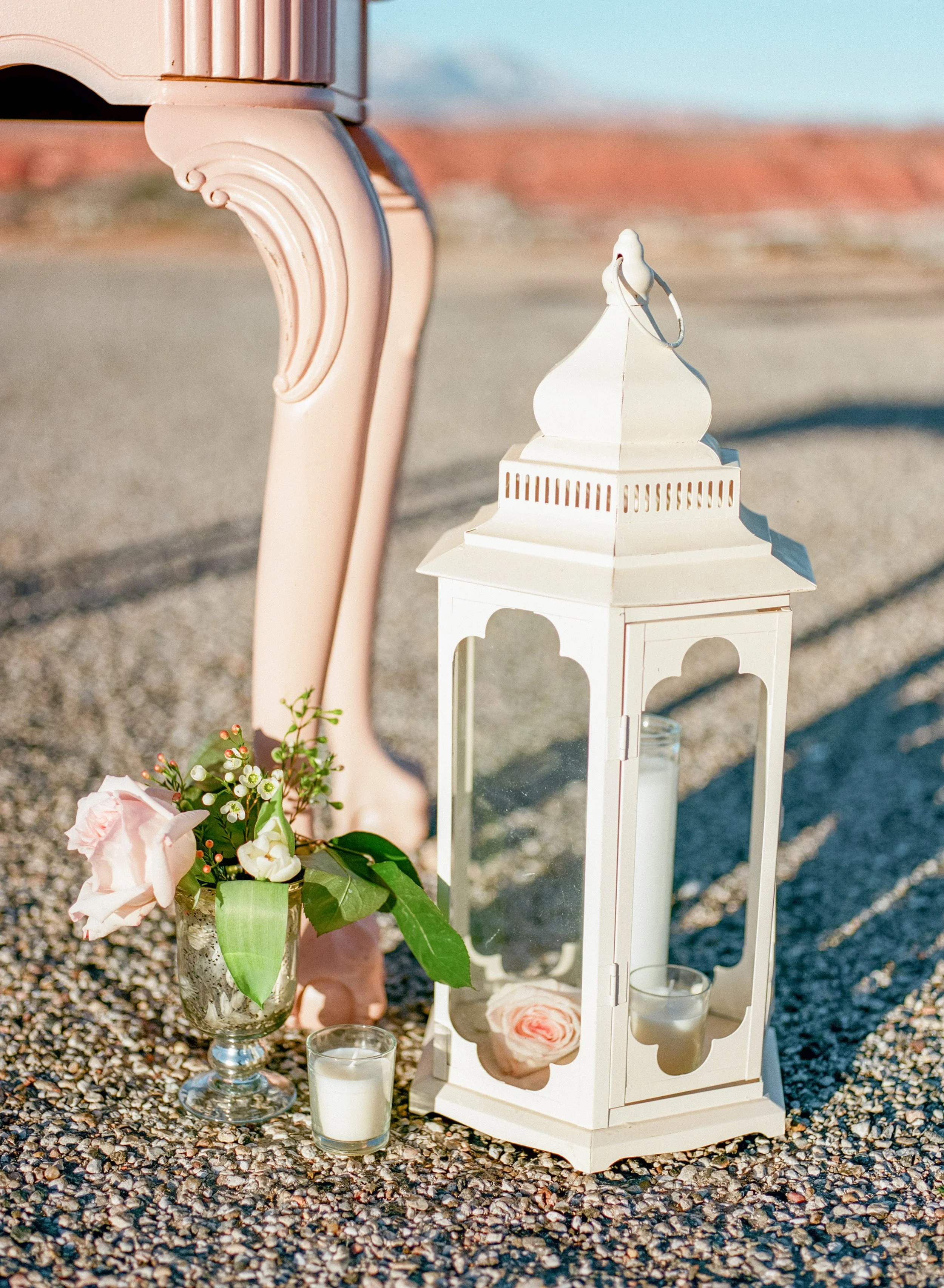A white lantern and candles, surrounded by pink flowers and greenery, placed on a gravel surface during daylight.