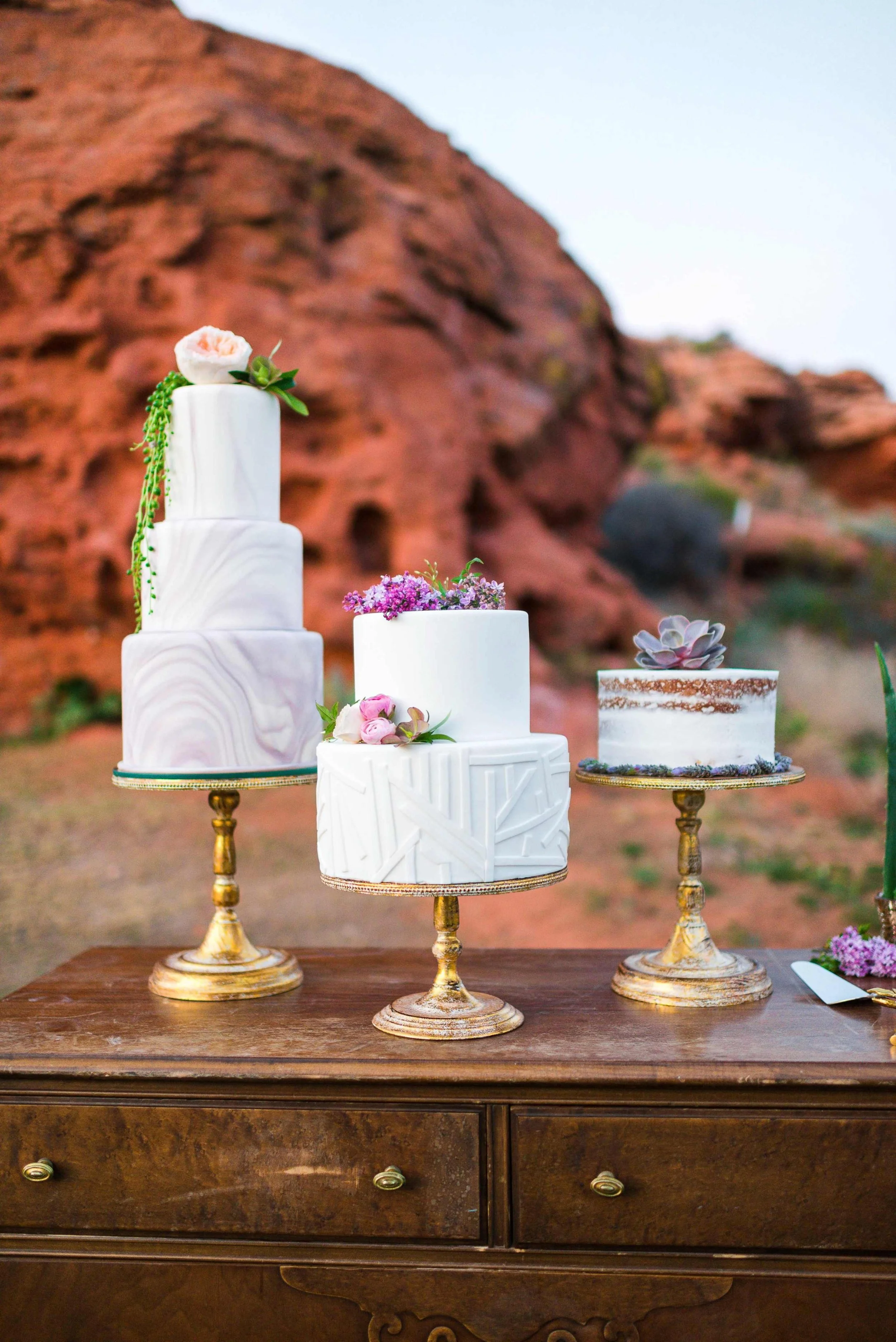 Three white decorative cakes on a wooden table with a red rock formation background. The cakes are adorned with flowers and greenery, each placed on a gold-colored cake stand.