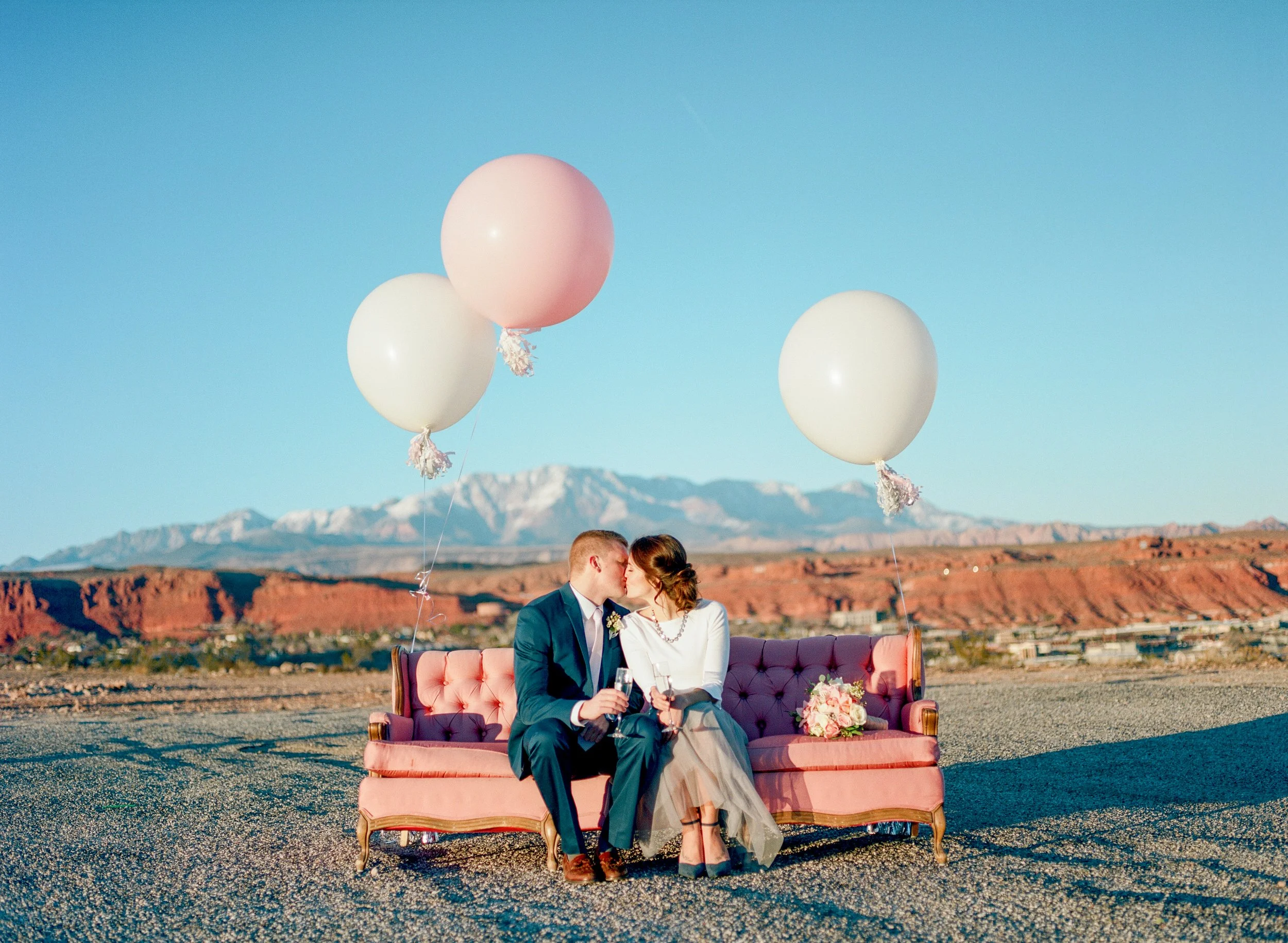A newlywed couple sitting on a pink sofa outdoors, kissing, with mountain ranges and a clear blue sky in the background. The bride is holding a bouquet of flowers, and there are three large balloons floating above them.