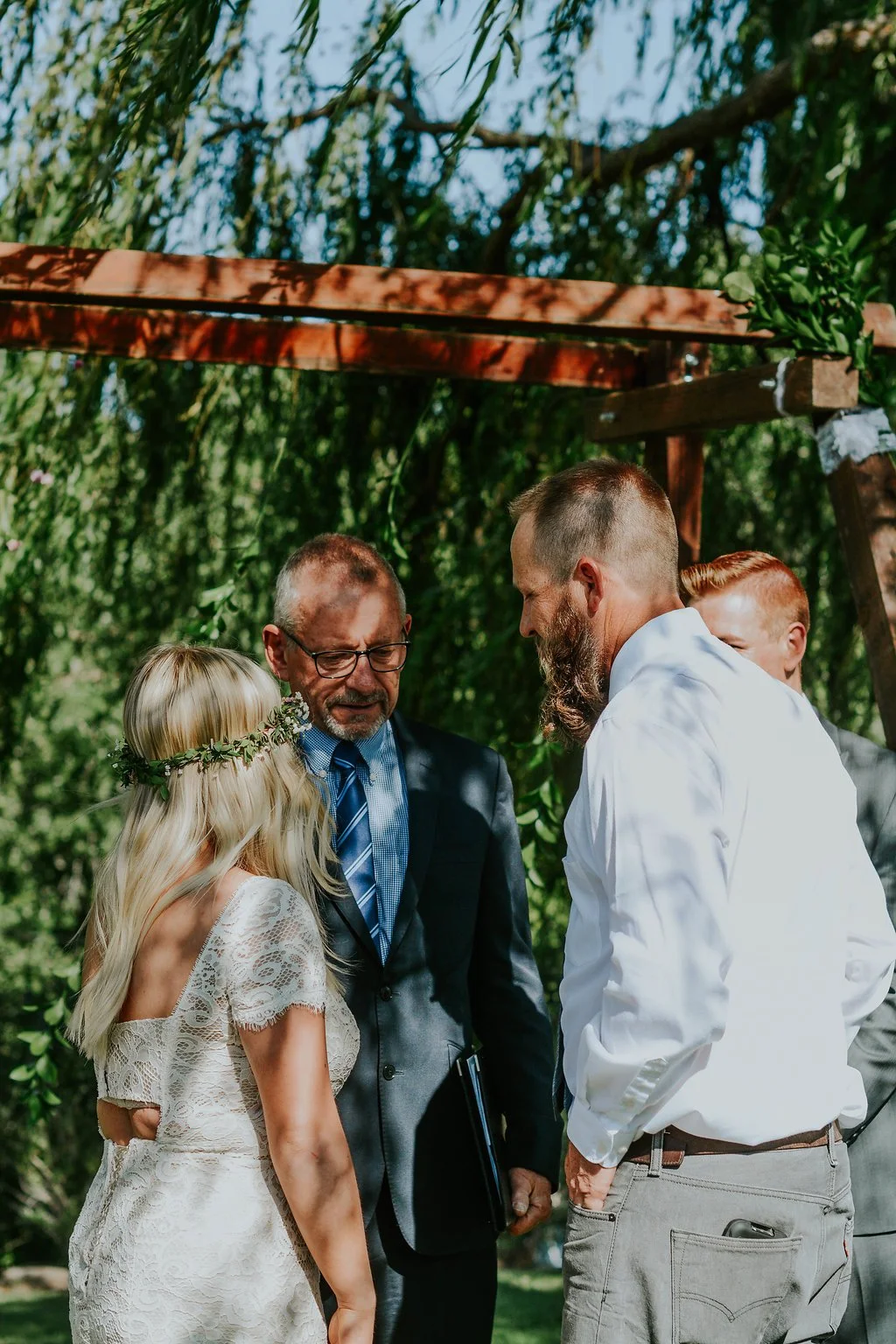 A couple having a wedding ceremony outdoors with officiant and groomsmen, under a wooden arch with greenery in the background.