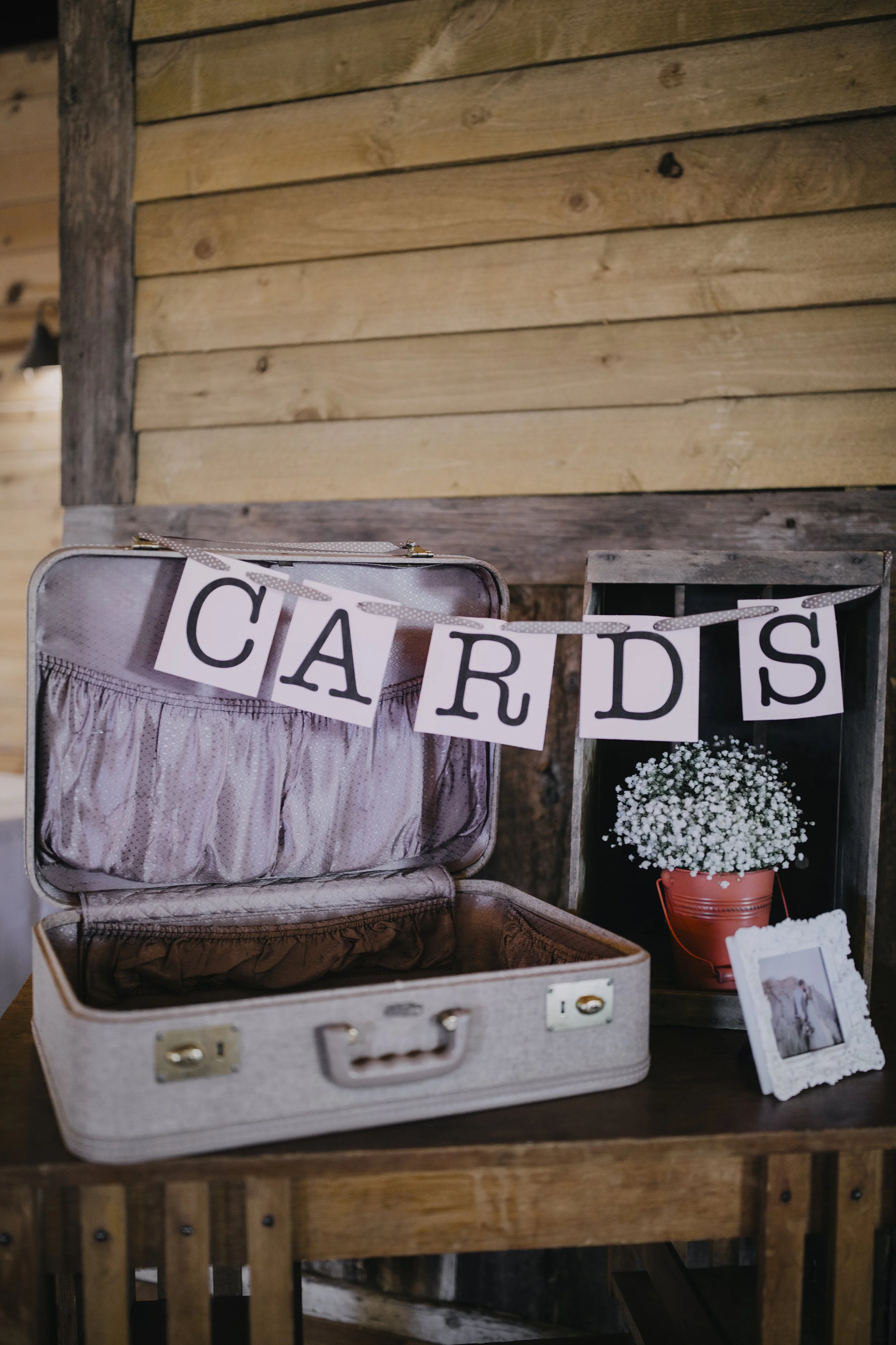 An open vintage pink suitcase on a wooden table, decorated for a card display, with a detectable label, and a small picture frame next to a potted plant with white flowers, set against a wooden wall background.