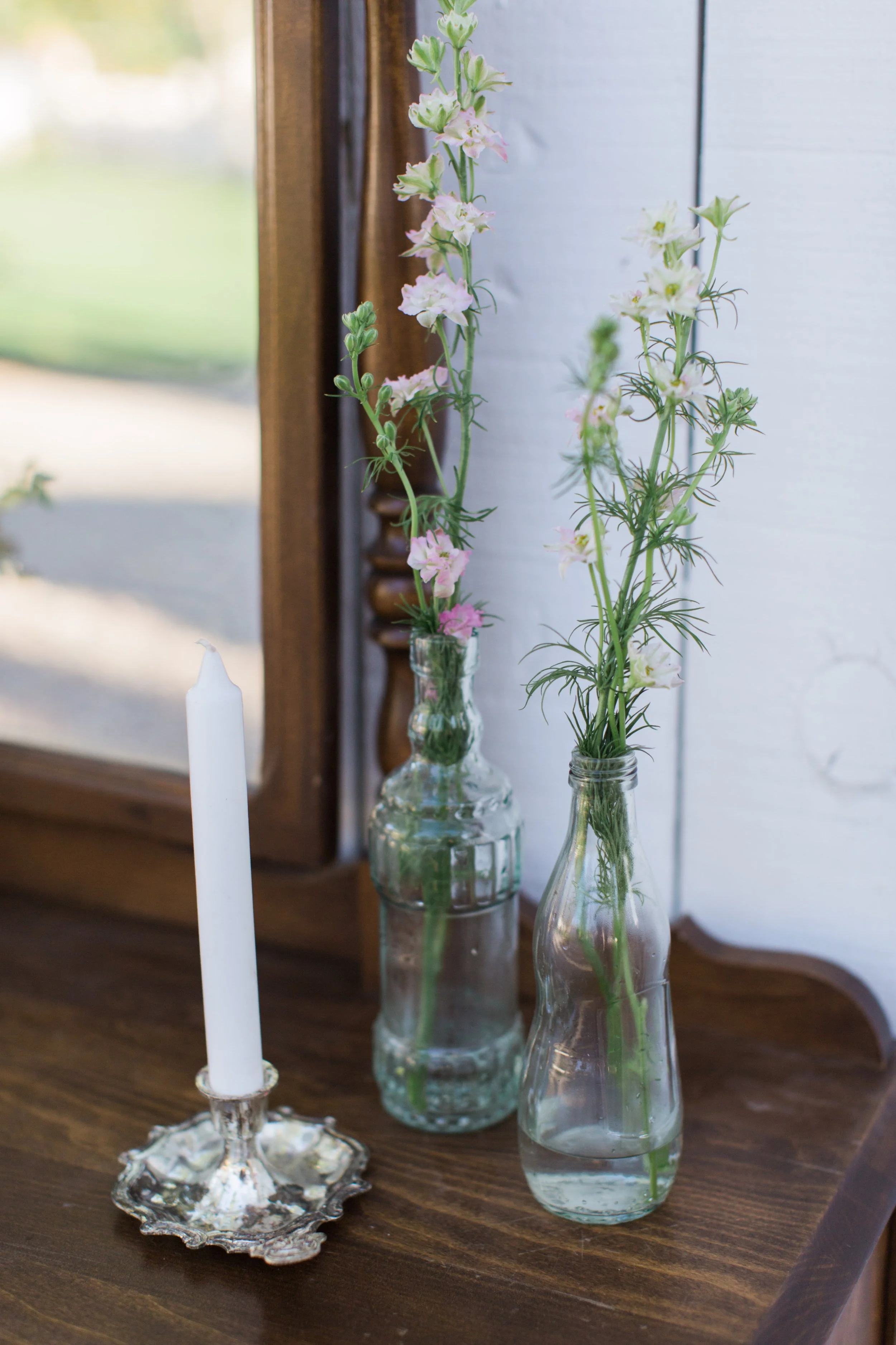 Three glass vases with tall, thin flowers placed on a brown wooden table, with a silver candlestick holder nearby.
