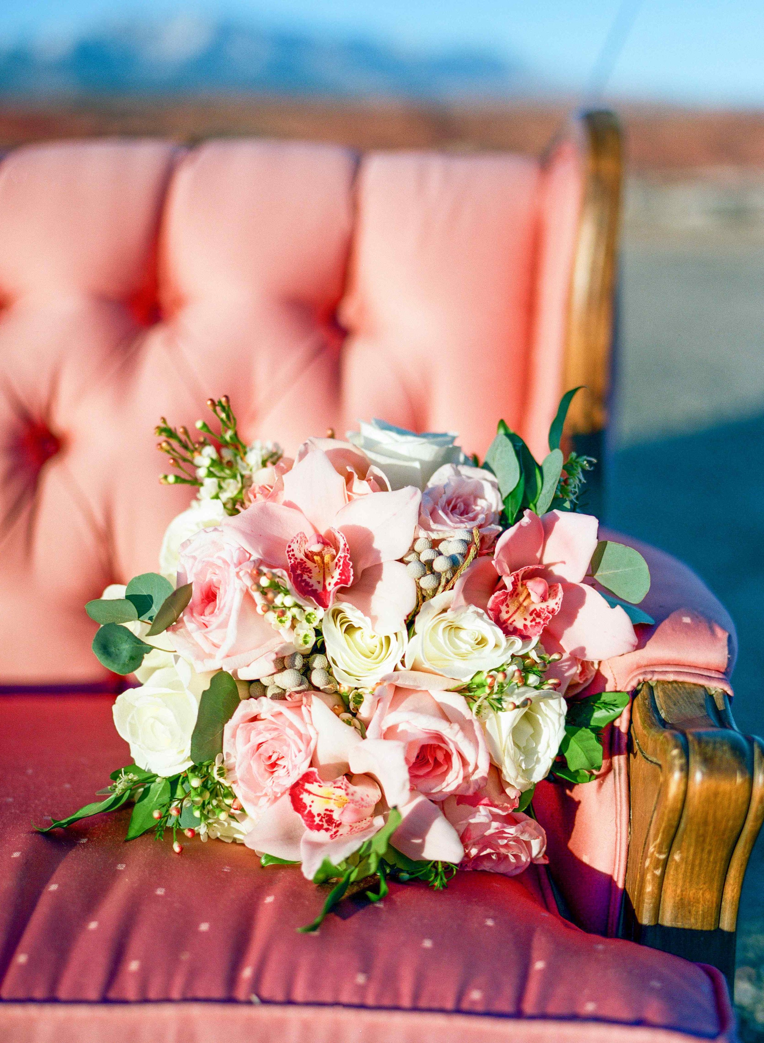 A floral bouquet with pink and white roses and greenery resting on a pink vintage upholstered bench with button tufting.