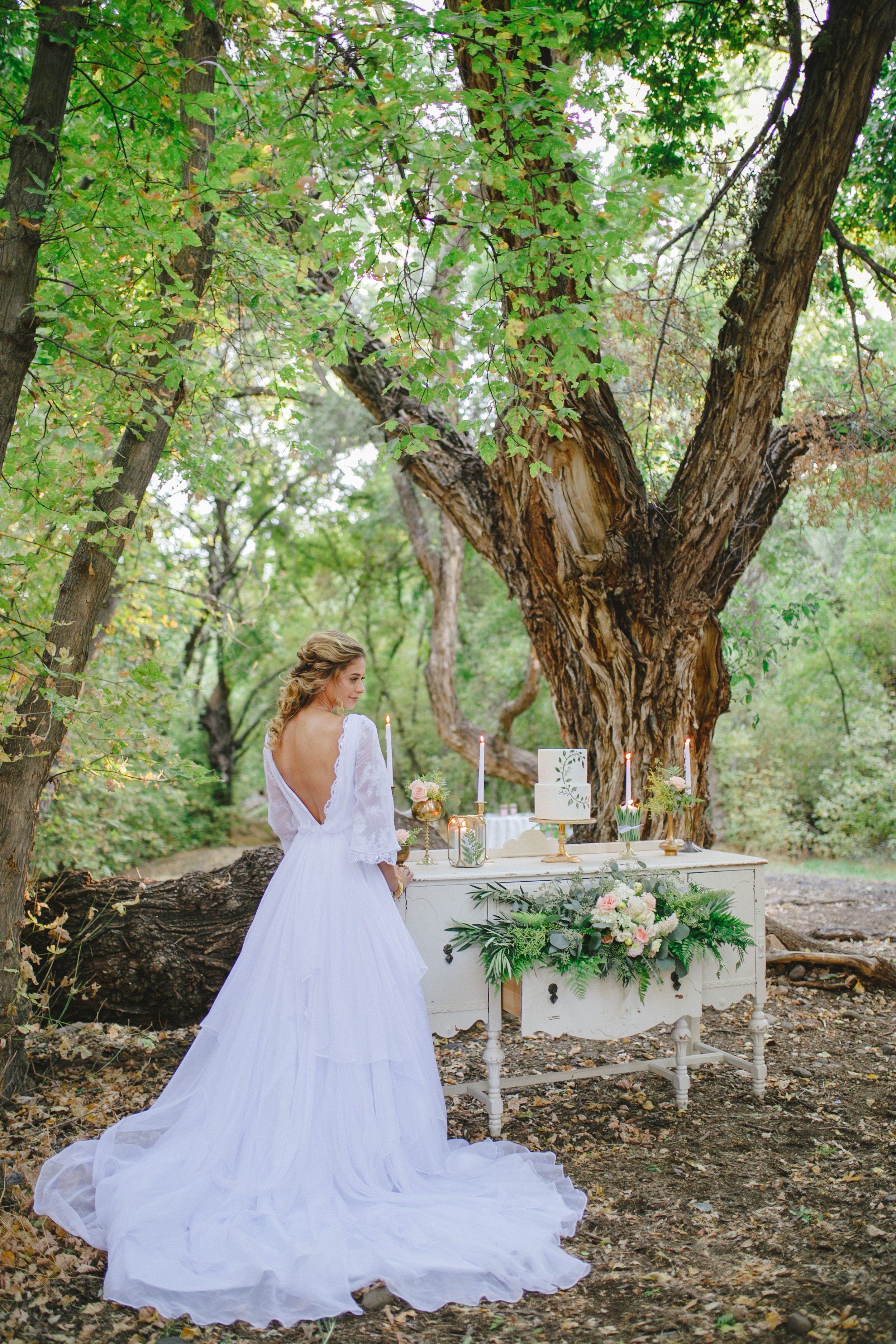 A woman in a white wedding dress standing outdoors in a forest, in front of a white stand with flowers and candles, under a large tree with green leaves.