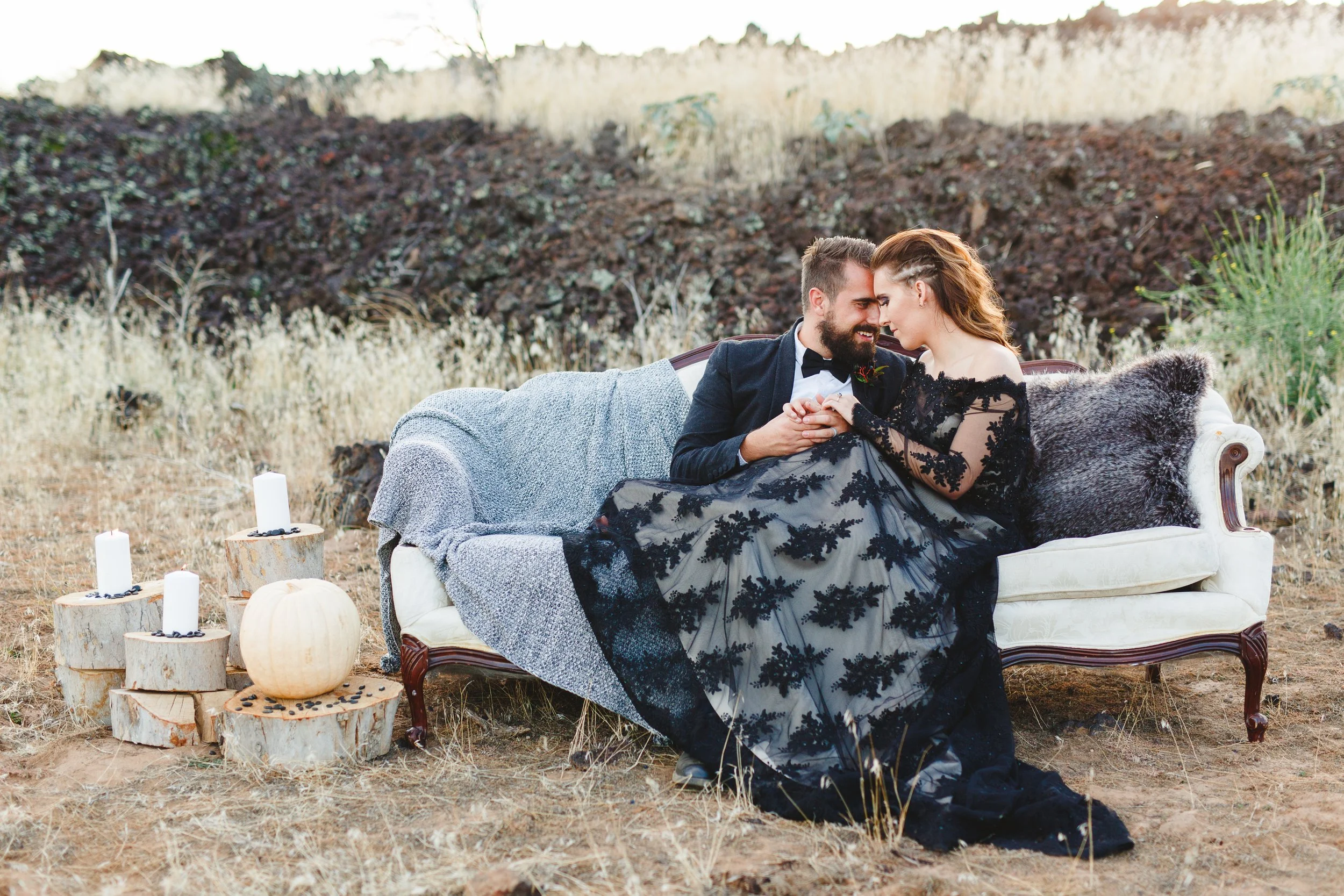 A couple sitting closely on a vintage sofa outdoors in a natural, rustic setting, with white candles and pumpkins around them.