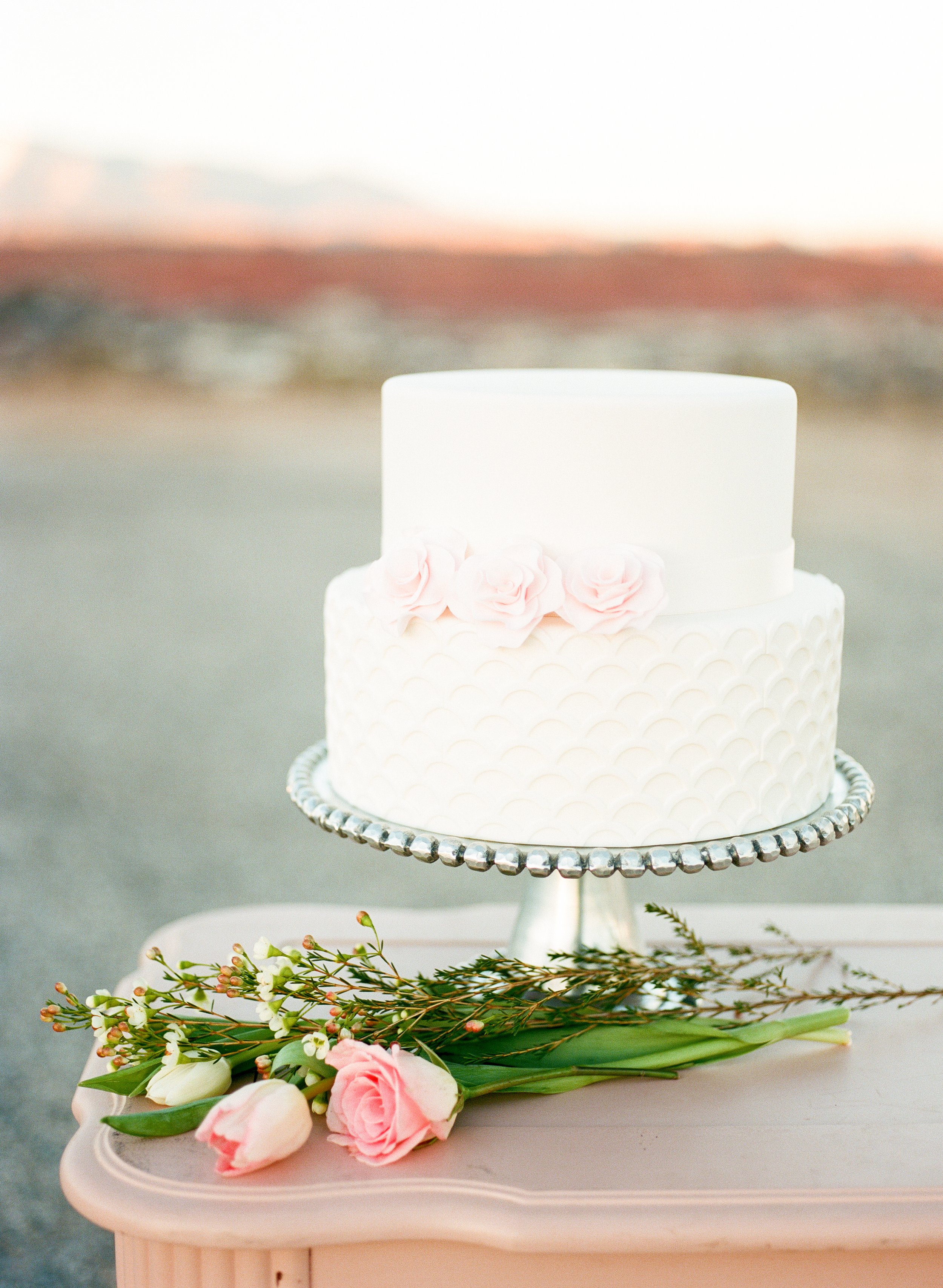 A white wedding cake decorated with pink roses on a silver cake stand, with floral arrangements of pink roses, white tulips, and greenery on a pink table in front.