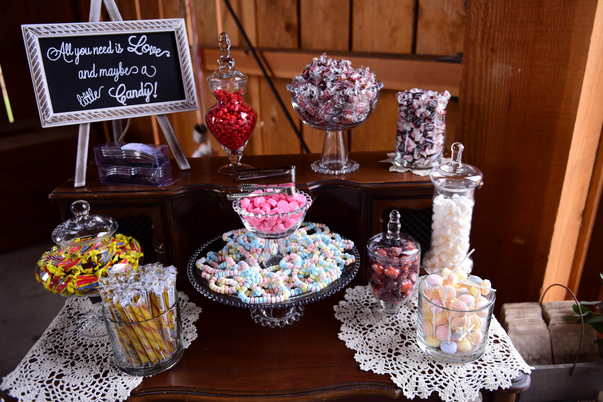 Assorted candies displayed on a table with a lace tablecloth, including jelly beans, lollipops, wrapped candies, and candy canes, with a decorative chalkboard sign that says, 'All you need is Love and maybe a little Candy!'