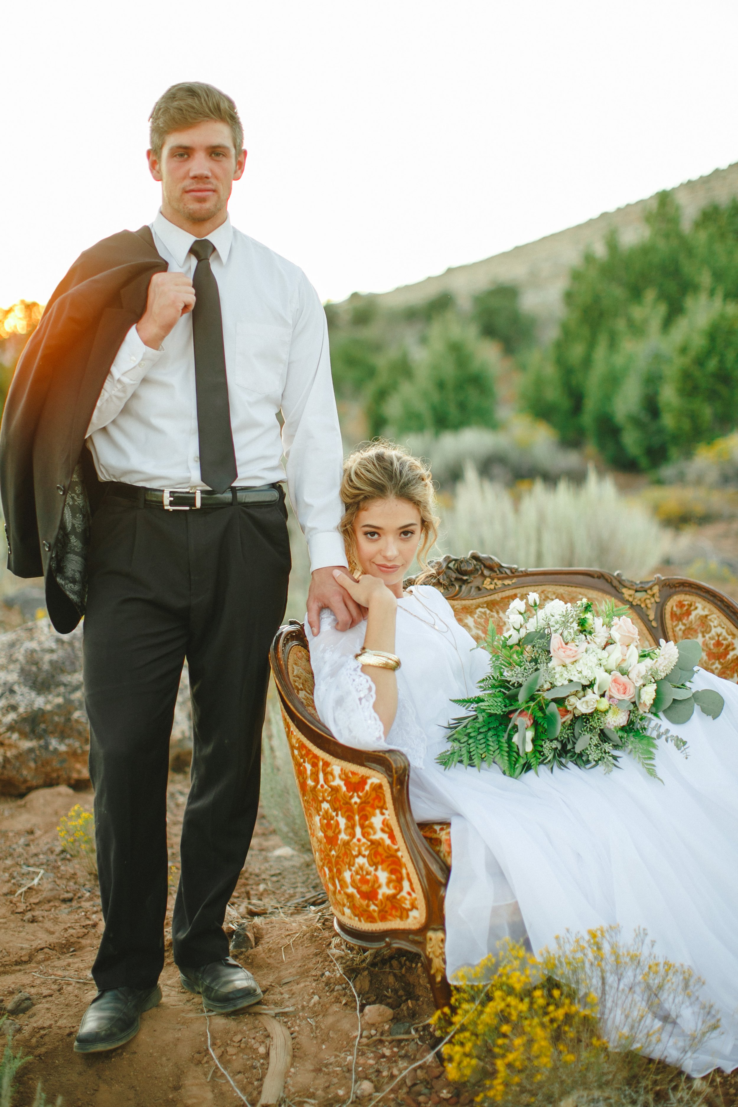 A young man in a white shirt, black tie, and black pants stands outdoors holding a black jacket over his shoulder. Next to him, a young woman in a white lace dress is seated on an ornate vintage sofa, holding a bouquet of flowers, with a scenic lands