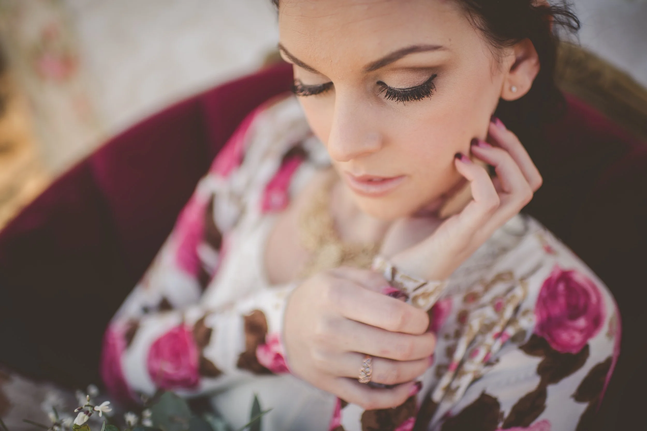 A woman with dark hair and makeup, wearing a floral top, looking down and touching her neck.