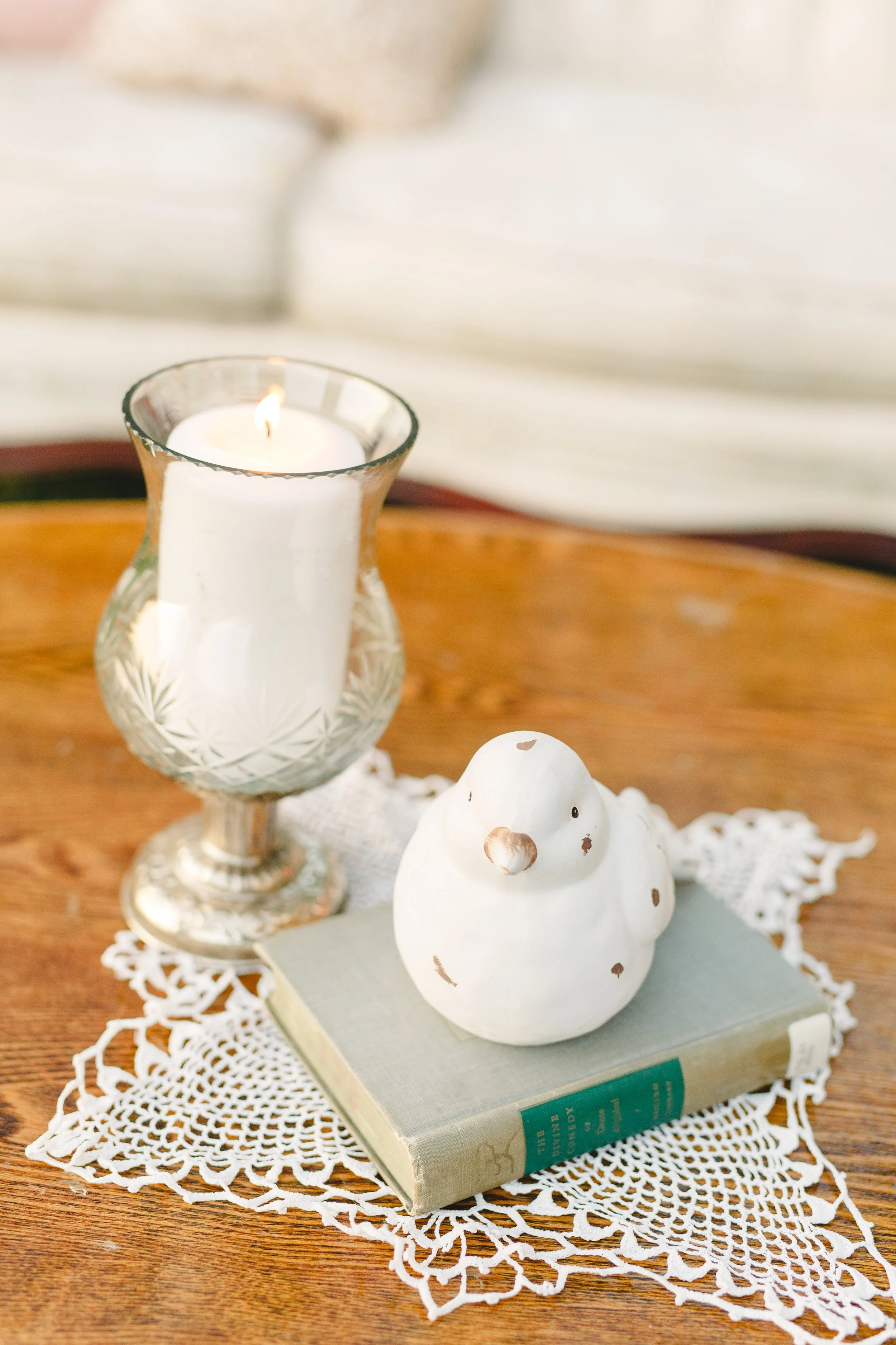 A white ceramic bird figurine on a book, placed on a decorative white lace doily, with a glass candle holder containing a lit white candle in the background on a wooden surface.