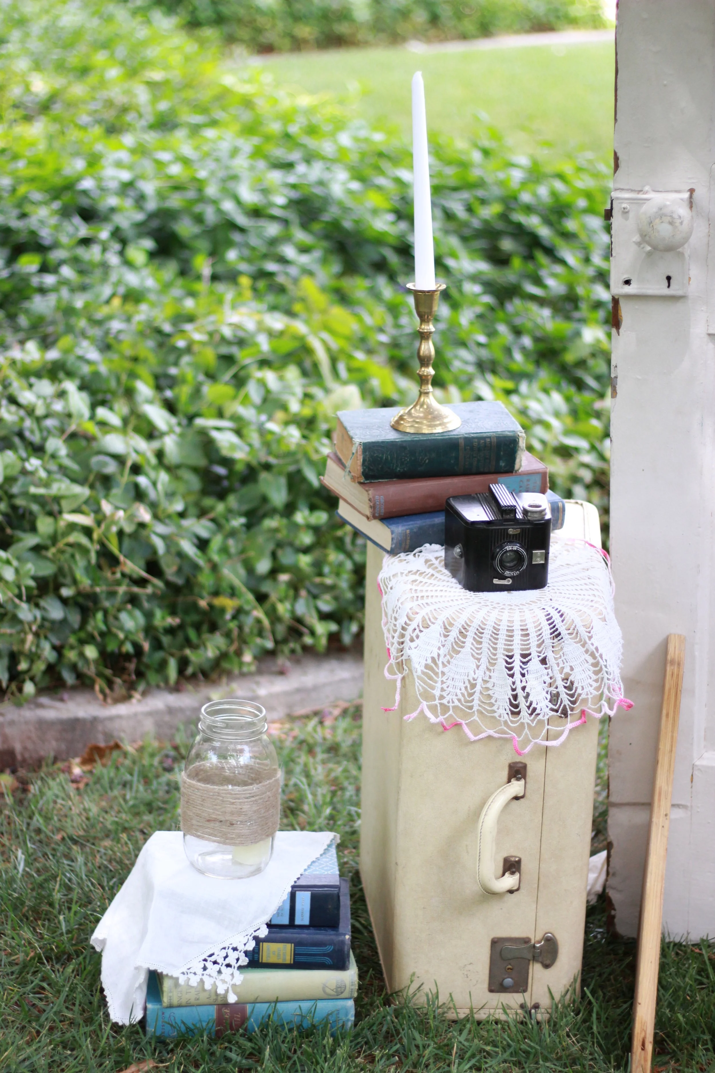 A rural outdoor scene with a white table covered in a lace tablecloth holding a glass jar and a row of books. Beside the table, there is a small stack of books on a stand with a lace cloth, a candle in a brass holder on top of some stacked books, and an old metal appliance or box with a paint-chipped white enamel finish and a vertical pipe. The background is filled with green shrubbery and grass.