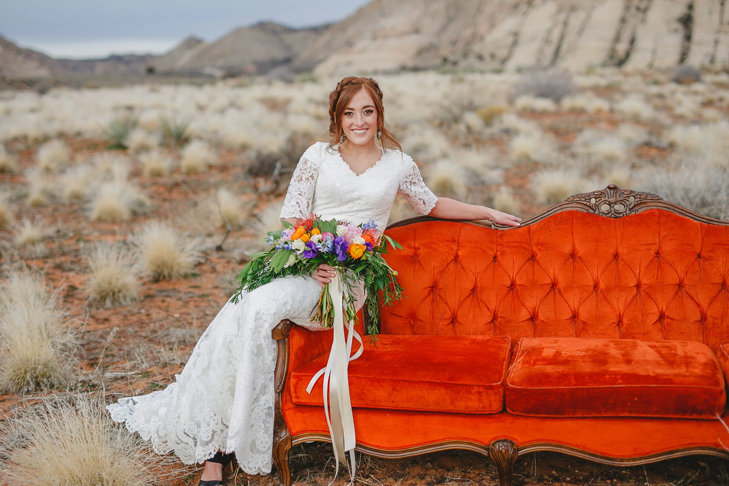 A woman in a white lace wedding dress holding a bouquet of colorful flowers, standing outdoors next to an orange vintage sofa in a desert landscape with mountains in the background.