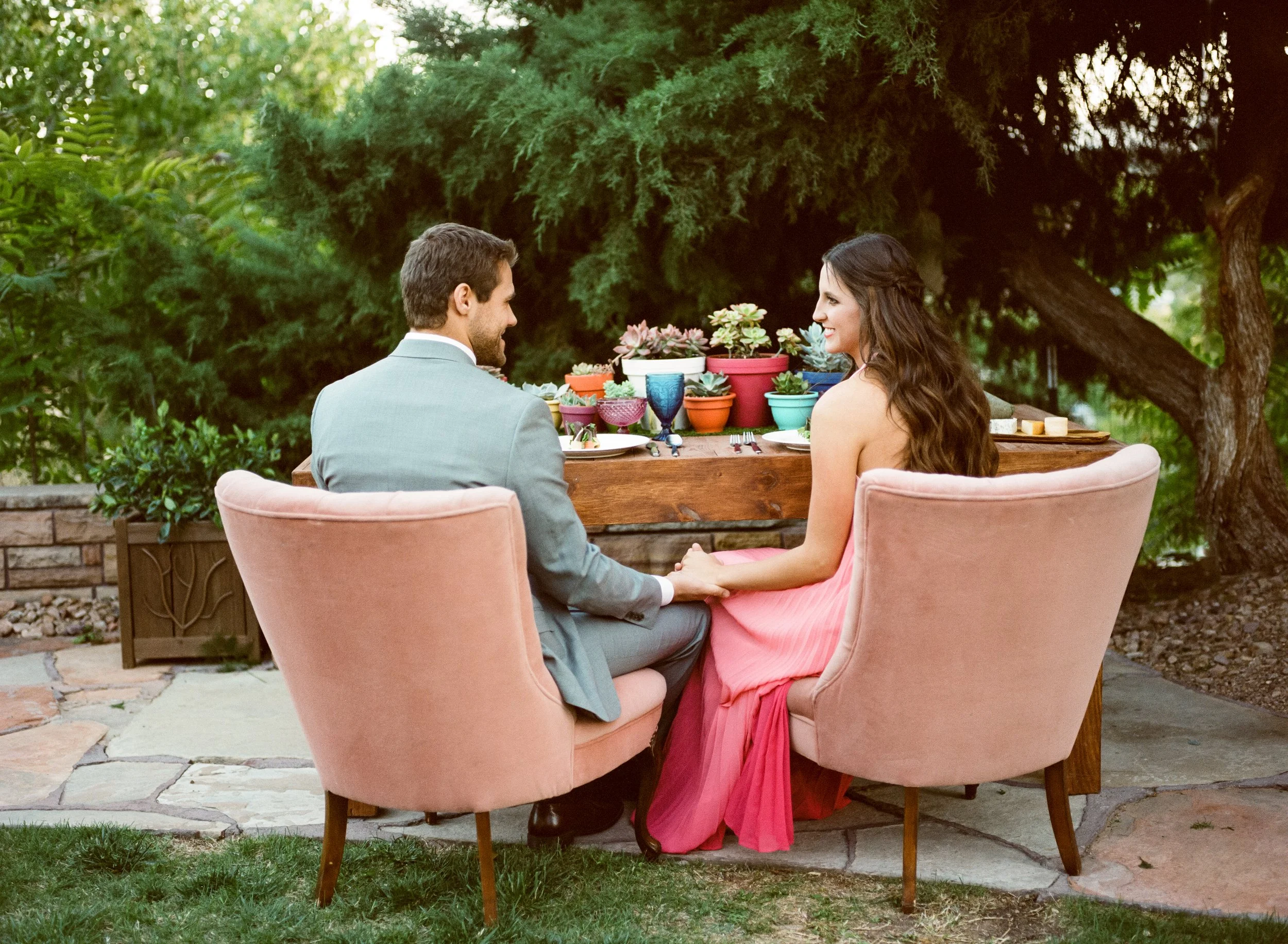 A couple is sitting at a table outdoors, holding hands and smiling at each other, with colorful potted plants and greenery in the background.