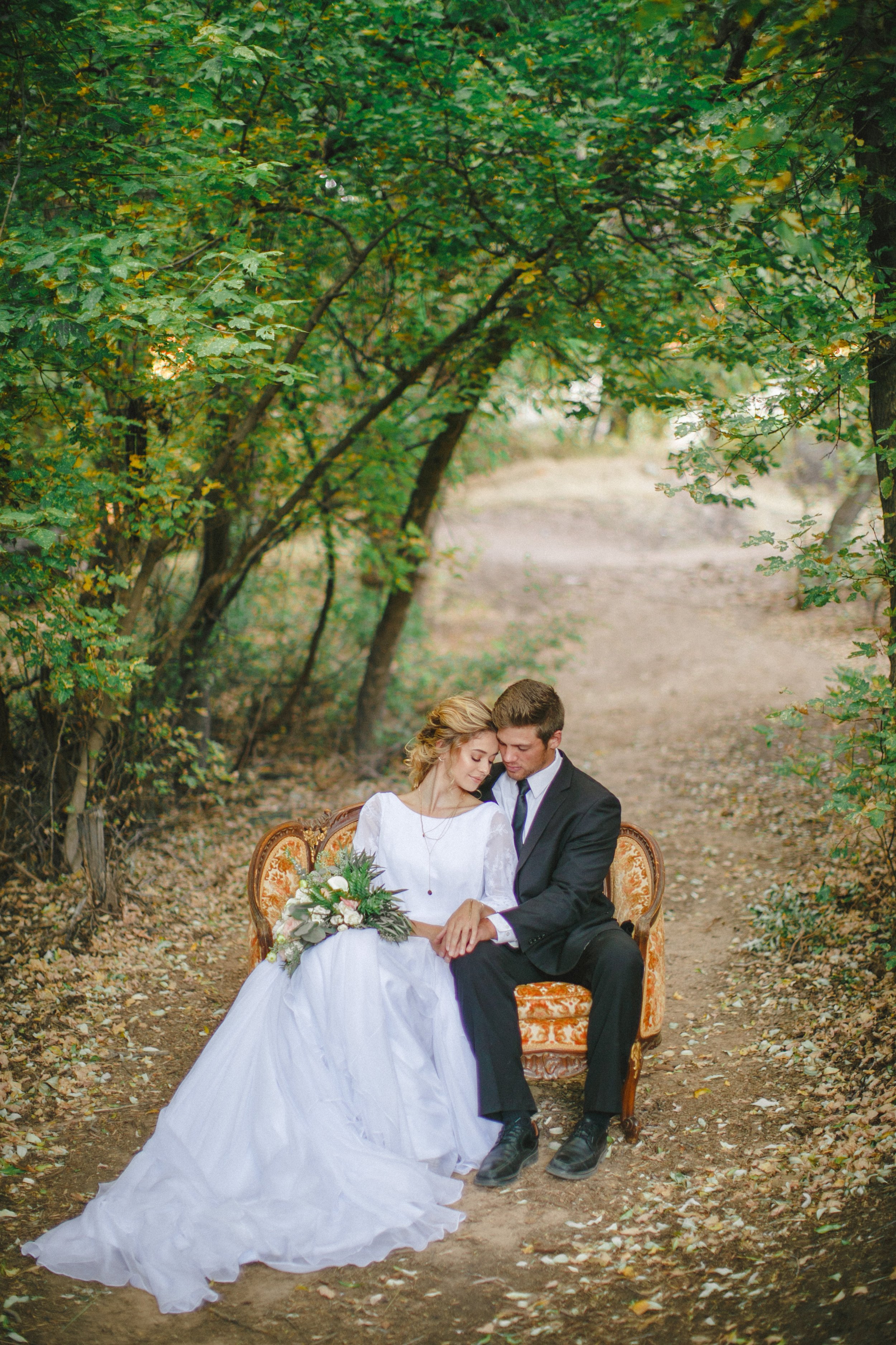 A couple dressed in wedding attire sitting on vintage orange and gold upholstered sofa in a forest with green trees and fallen leaves, they are leaning close with eyes closed, holding hands and a bouquet of flowers