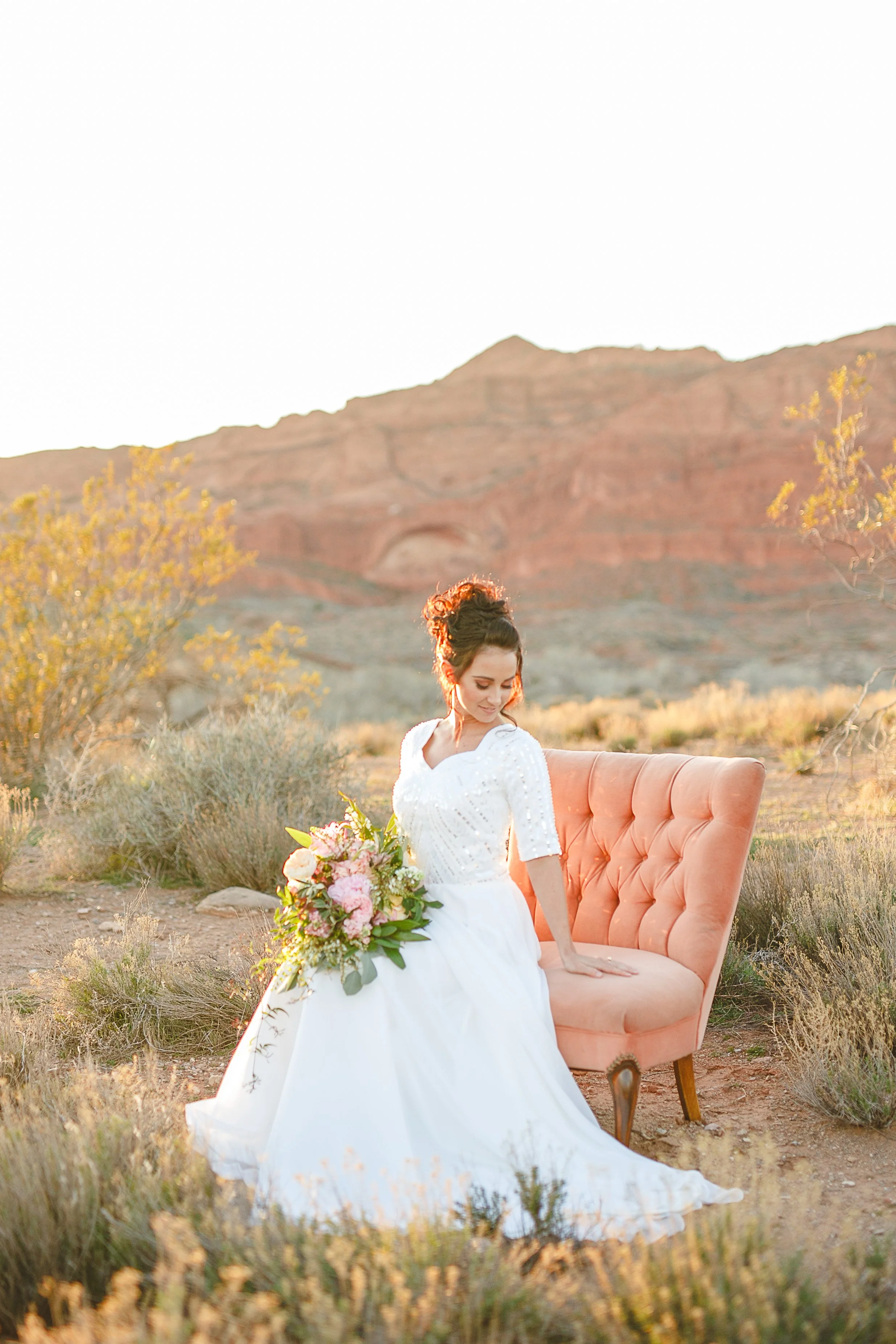 A woman in a white wedding dress sitting on a pink vintage sofa in a desert landscape with mountains and trees, holding a bouquet of pink and white flowers, with the sun setting.