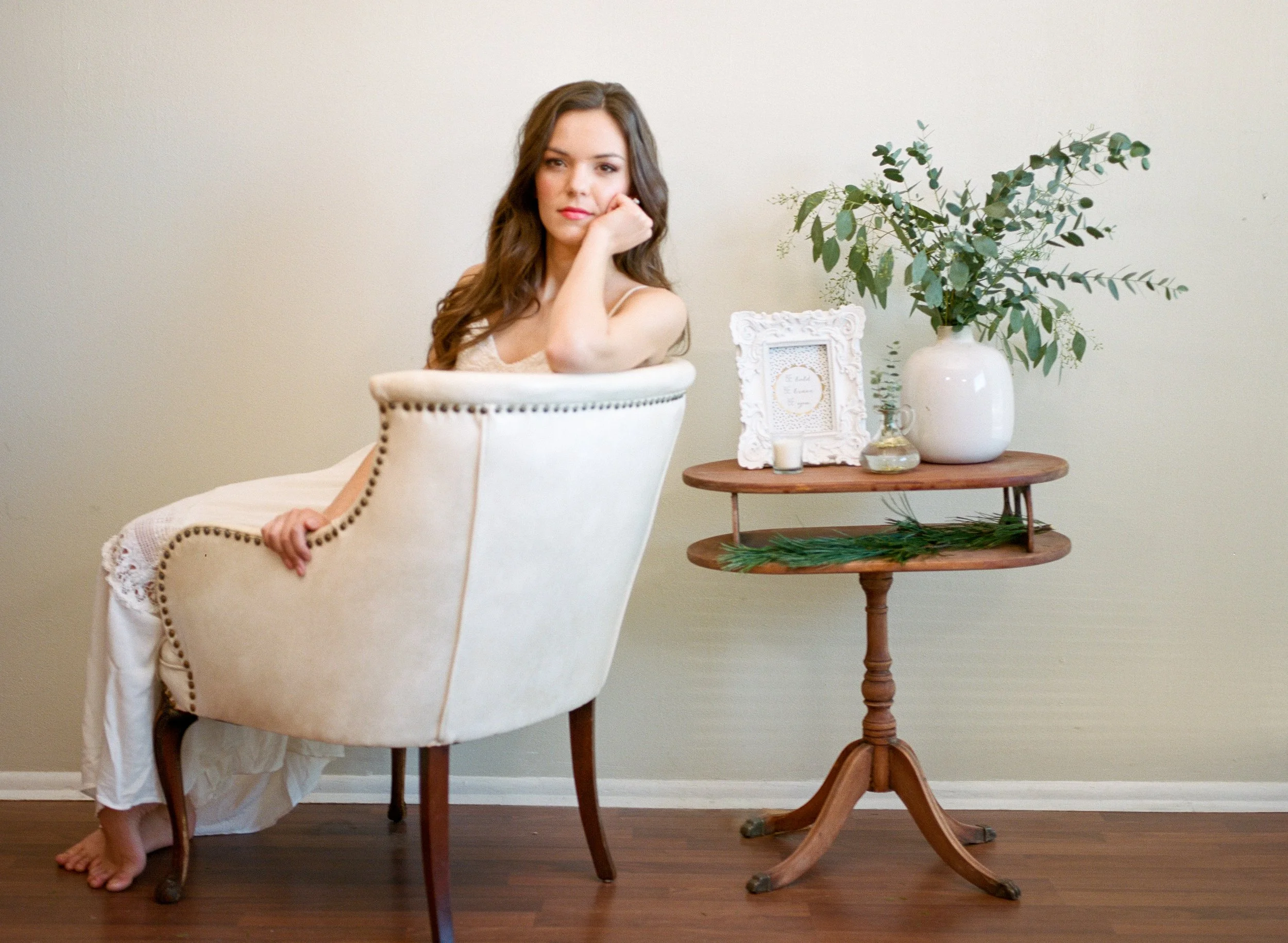 A young woman with long brown hair sitting in a cream-colored armchair with nailhead trim, resting her head on her hand. She is on a wooden floor next to a small round wooden table with a white vase of greenery and a decorative picture frame. The background features a plain, light-colored wall.