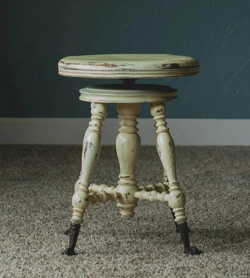 An antique white wooden stool with turned legs and black metal feet, sitting on a beige carpet in front of a dark blue wall.