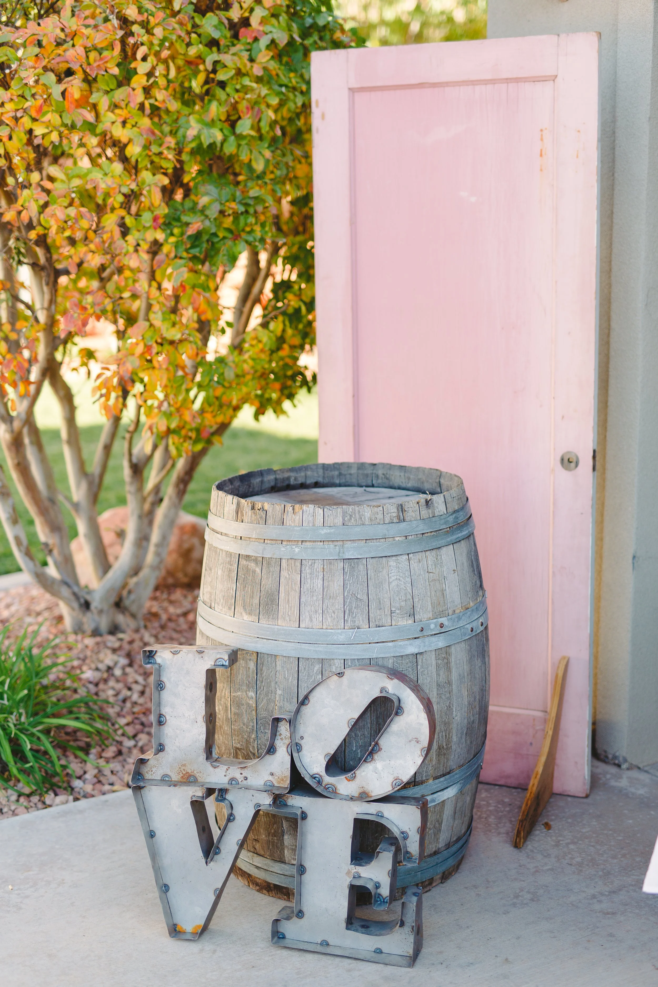 A wooden barrel with metal bands and a metal 'LOVE' sign in front of it, placed outdoors near a pink door and a tree with green and orange leaves.