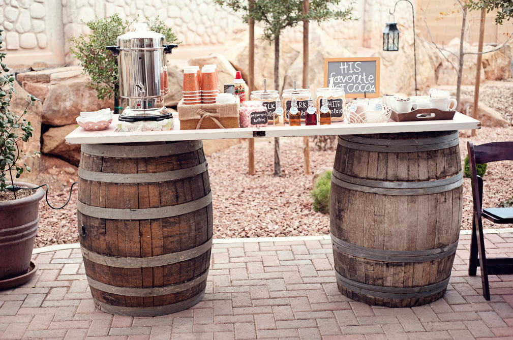 Outdoor coffee station set up on two wooden barrel tables with coffee cups, syrup bottles, and a chalkboard sign that reads "His favorite cocoa."