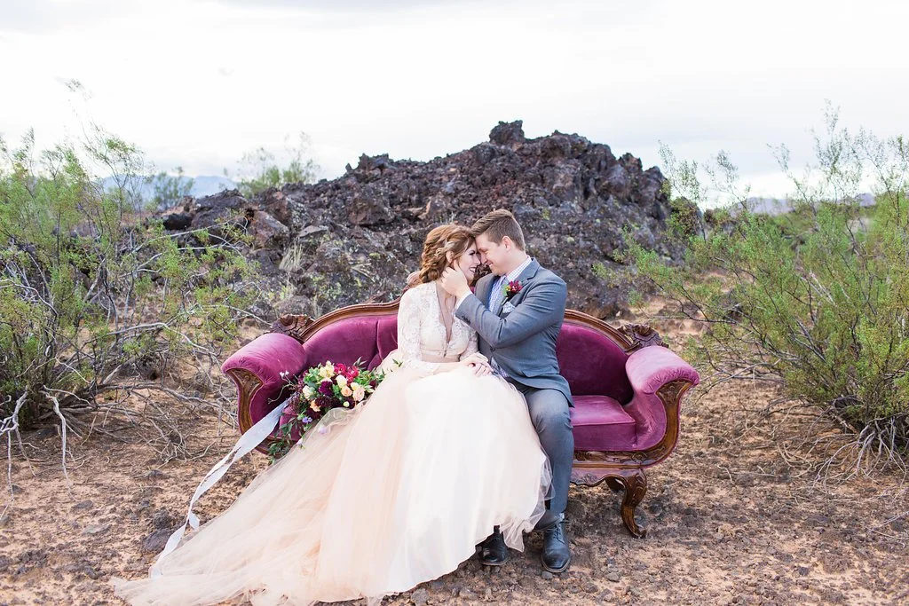 A bride and groom sit closely on a vintage purple sofa in a desert landscape, with rocks and sparse shrubs in the background. The bride wears a white wedding gown, and the groom wears a gray suit. They are leaning into each other, smiling, with a bouquet of flowers beside them.