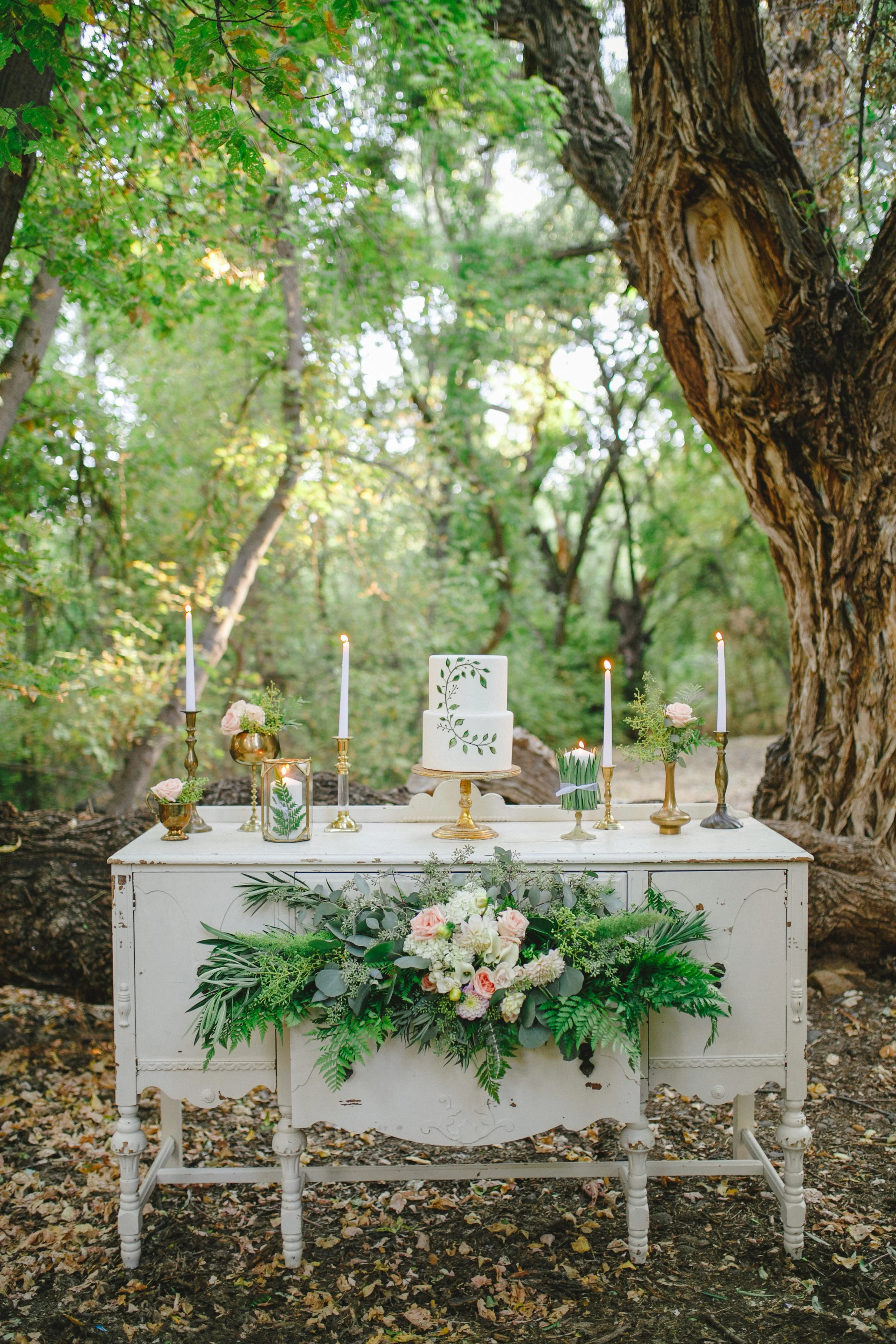 A white vintage-style dresser decorated with a flower arrangement at the bottom and adorned with a two-tier white cake decorated with green leafy vines. Surrounding the cake are lit candles in gold candle holders, some with pink roses. The scene is set outdoors in a wooded area with green foliage and a large tree trunk in the background, suggesting a rustic or garden wedding celebration.