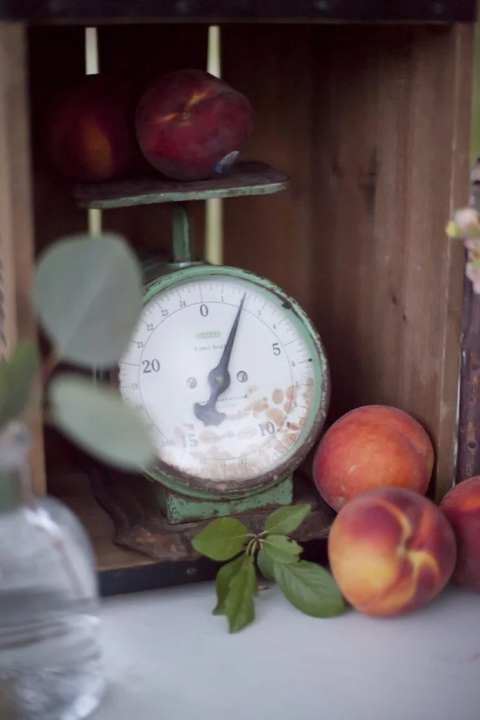 Vintage green kitchen scale with peaches on an open wooden shelf, some peaches resting against the scale, green leaves in the foreground, and a glass container on the left.