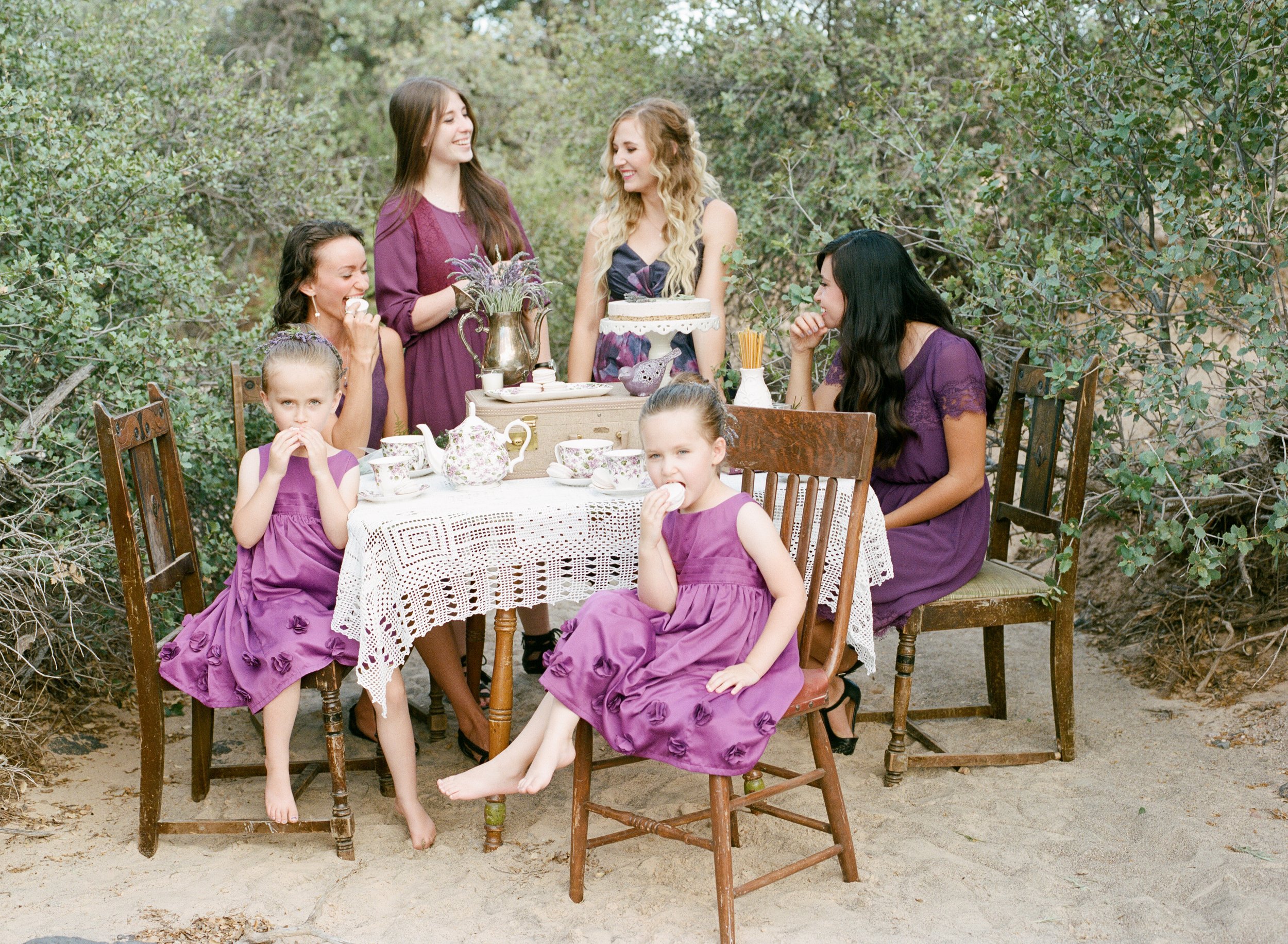 A group of women and young girls celebrating a birthday outdoors, with a table set with teapots and cups, and a cake in the center, surrounded by green foliage.