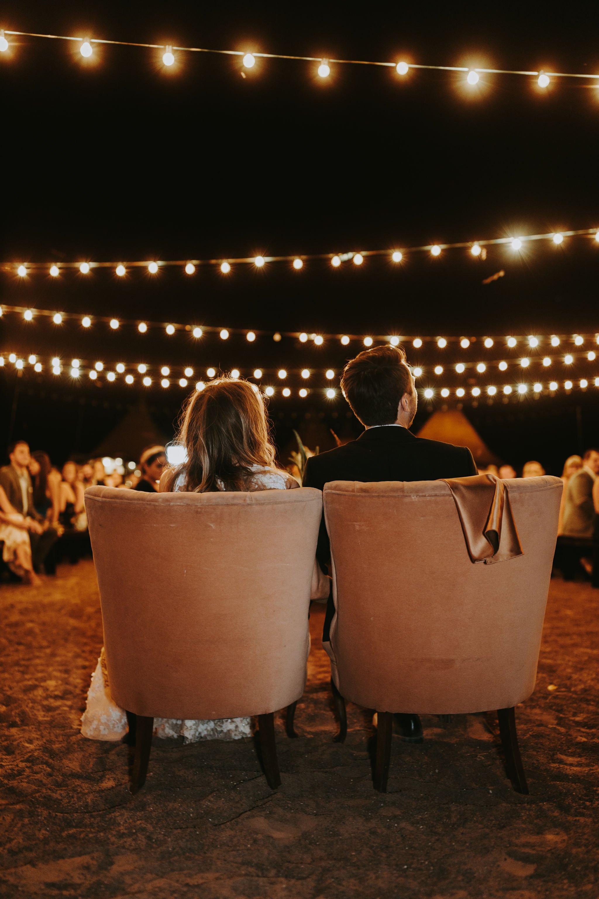A couple sitting on chairs at an outdoor event at night, decorated with string lights overhead, with a crowd in the background.