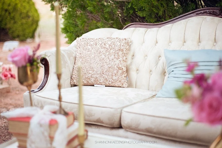 A vintage white tufted sofa with dark wood trim, two decorative pillows (one pink, one blue), and greenery in the background, outdoors.