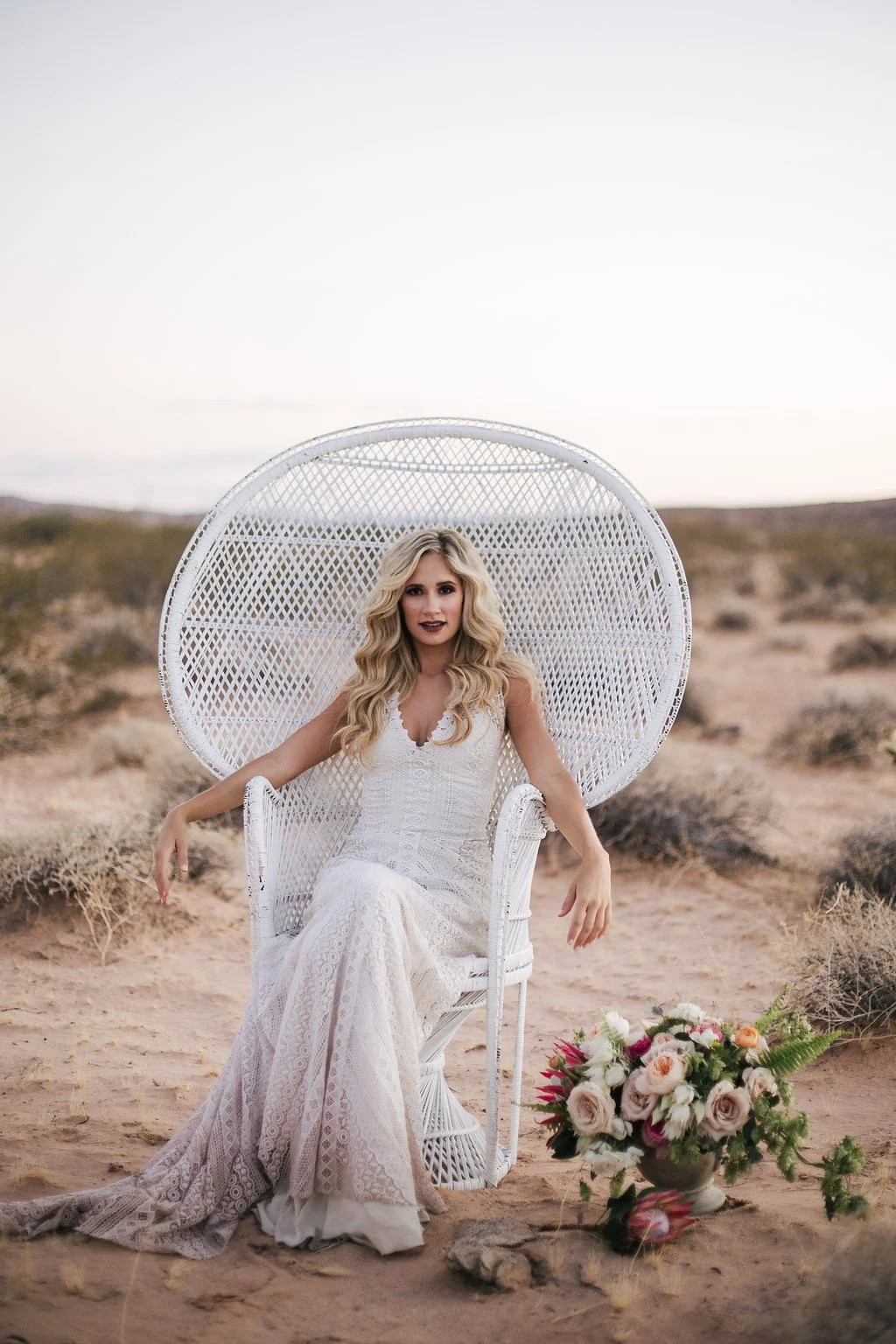 A woman in a white lace dress sitting in a white peacock chair outdoors in a desert landscape with a bouquet of pink and white flowers on the ground nearby.