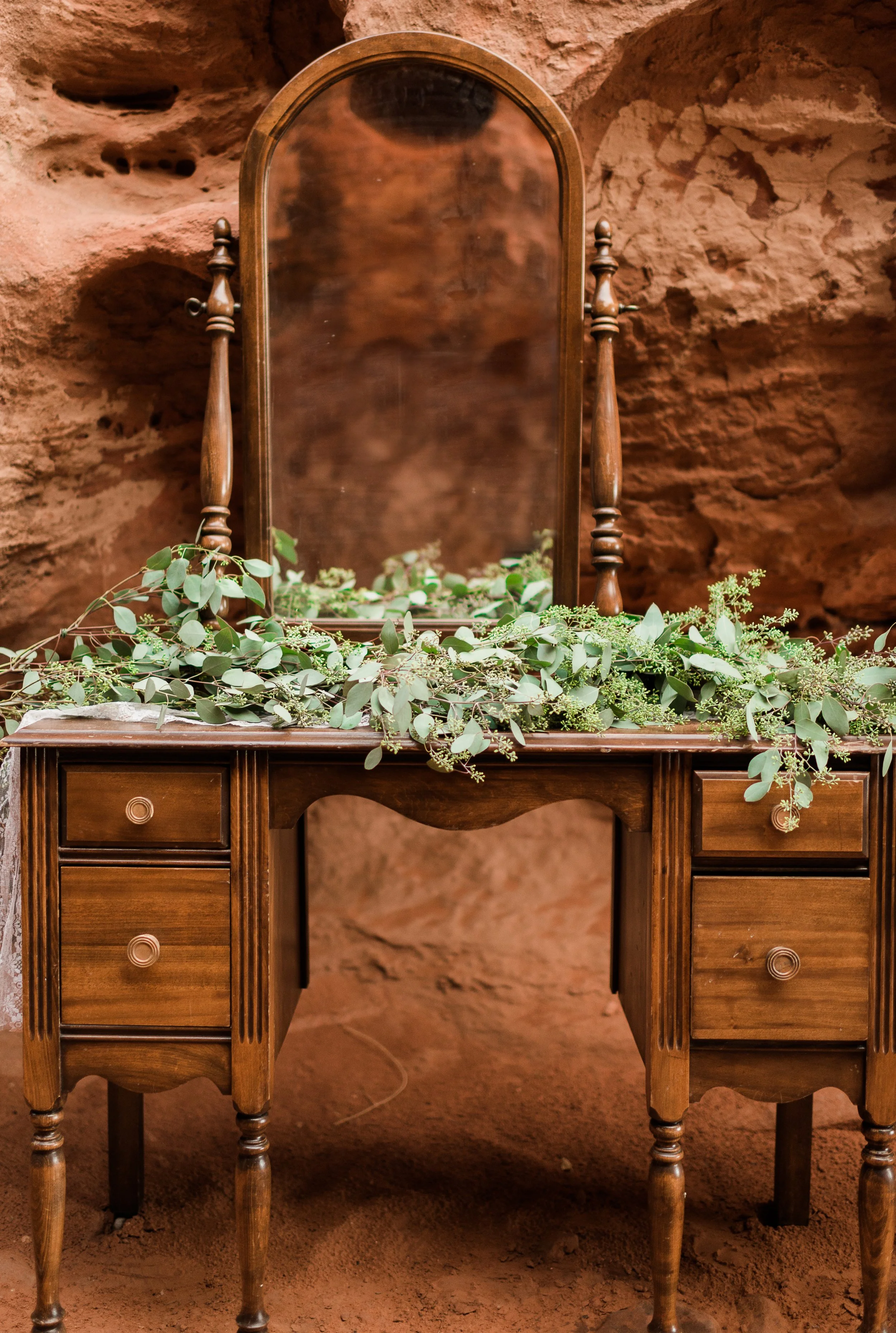 A vintage wooden vanity table with a mirror, decorated with green foliage, against a brown textured wall.