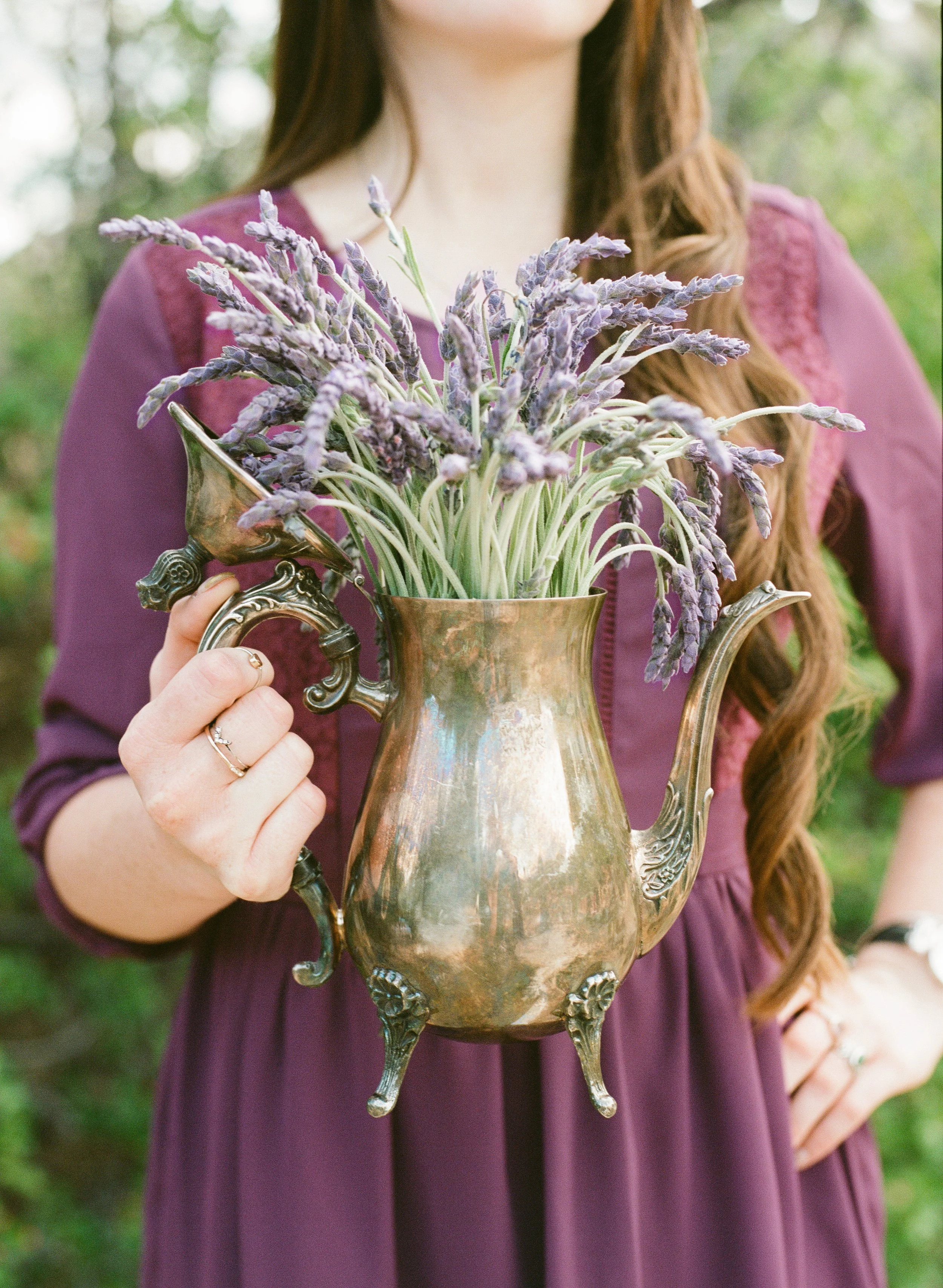 A woman with long wavy brown hair wearing a purple dress holding an ornate brass pitcher filled with lavender flowers outdoors.