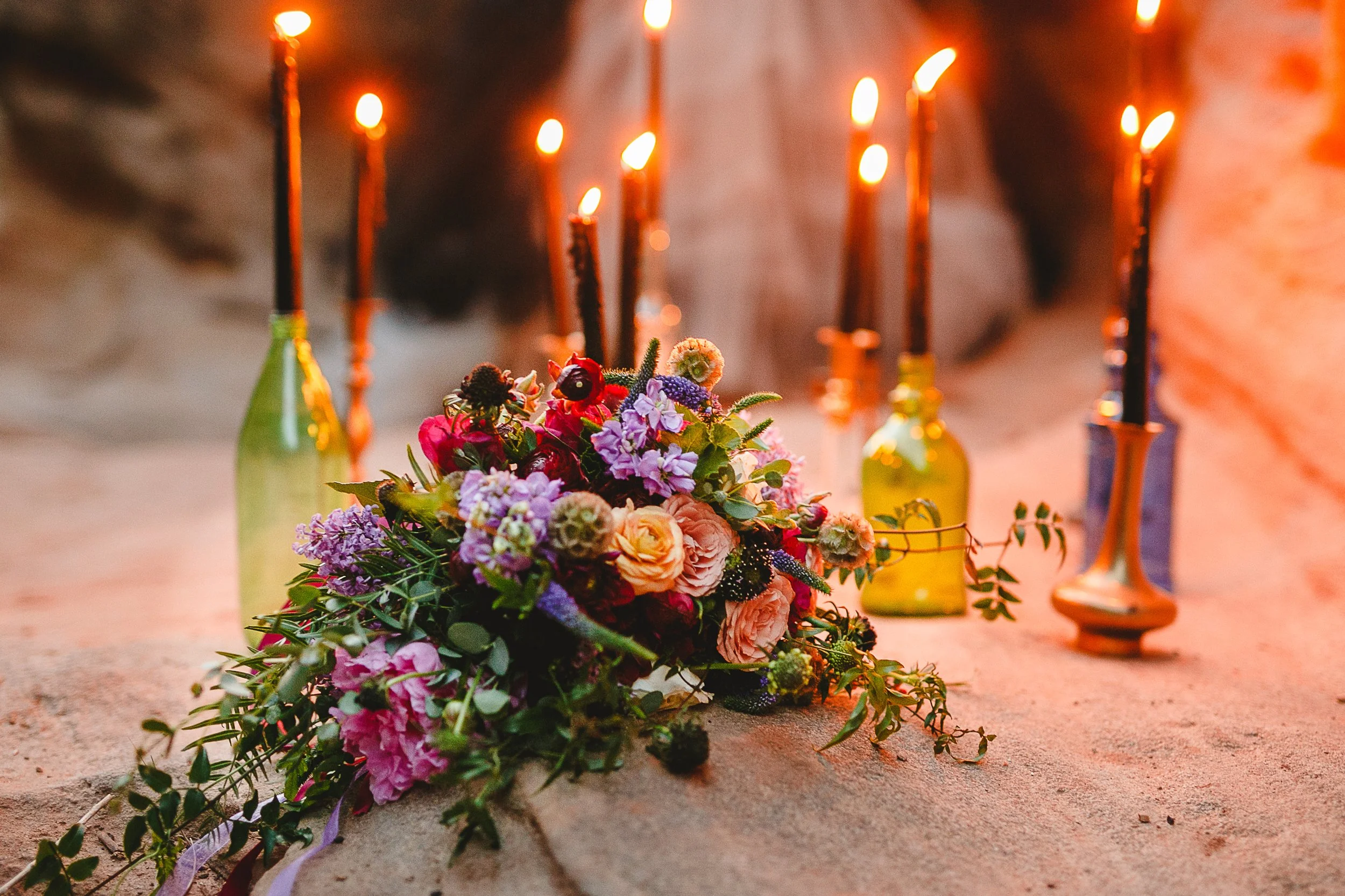 Close-up of a colorful flower arrangement on the ground with lit candles in bottles in the background.