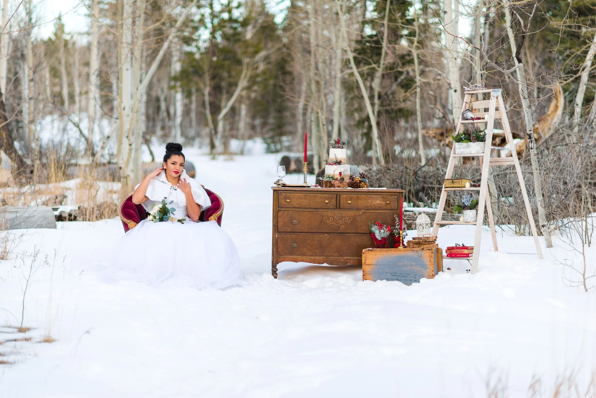 A woman sitting on a red velvet chair in a snowy outdoor setting, decorated with holiday items including a cake, candles, and festive ornaments, with a wooden ladder displaying plant arrangements and ornaments nearby.