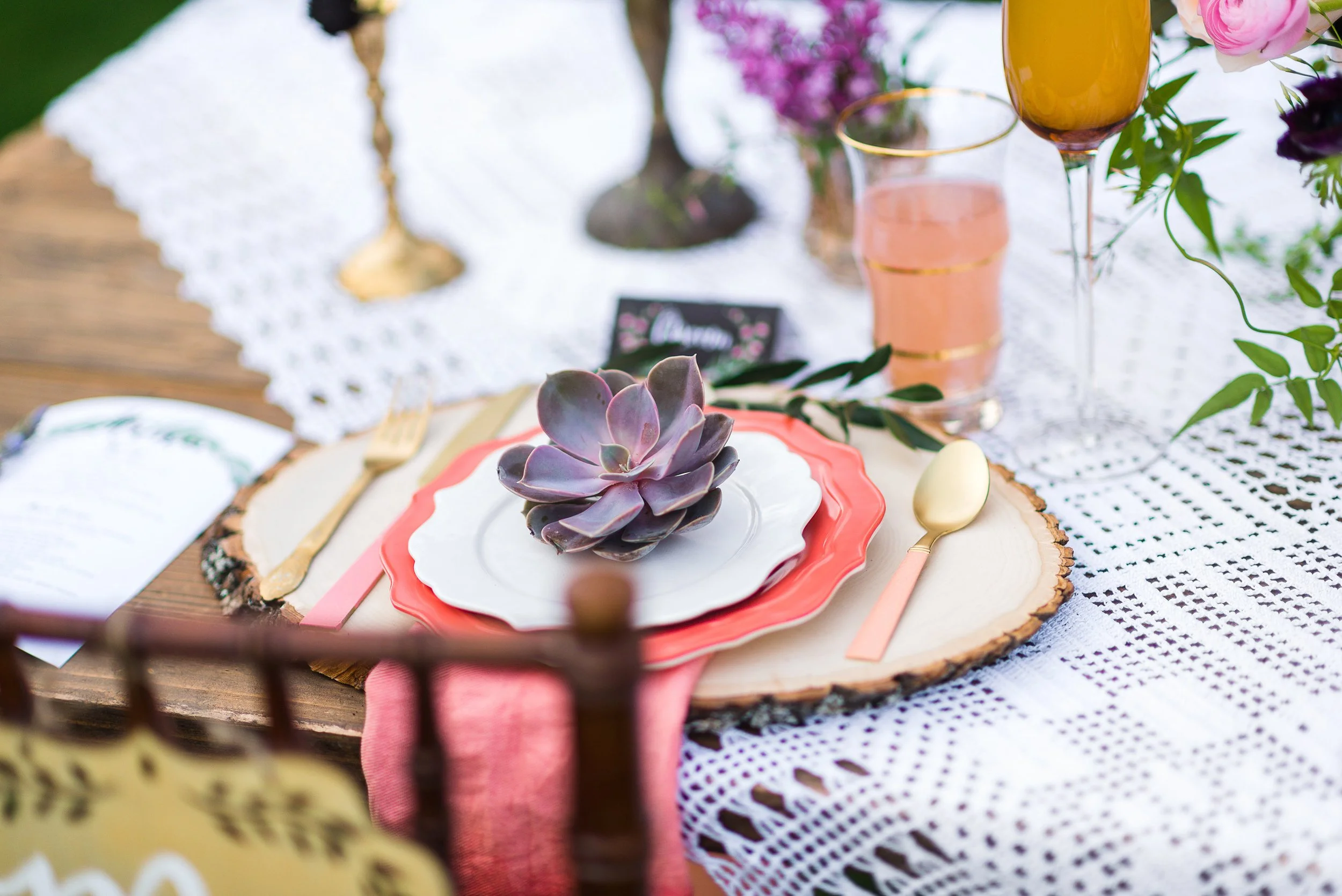 A close-up of a decorated table with a purple succulent on a pink and white plate, set on a wooden table with a white lace tablecloth, pink cutlery, and pink and purple flowers in the background.