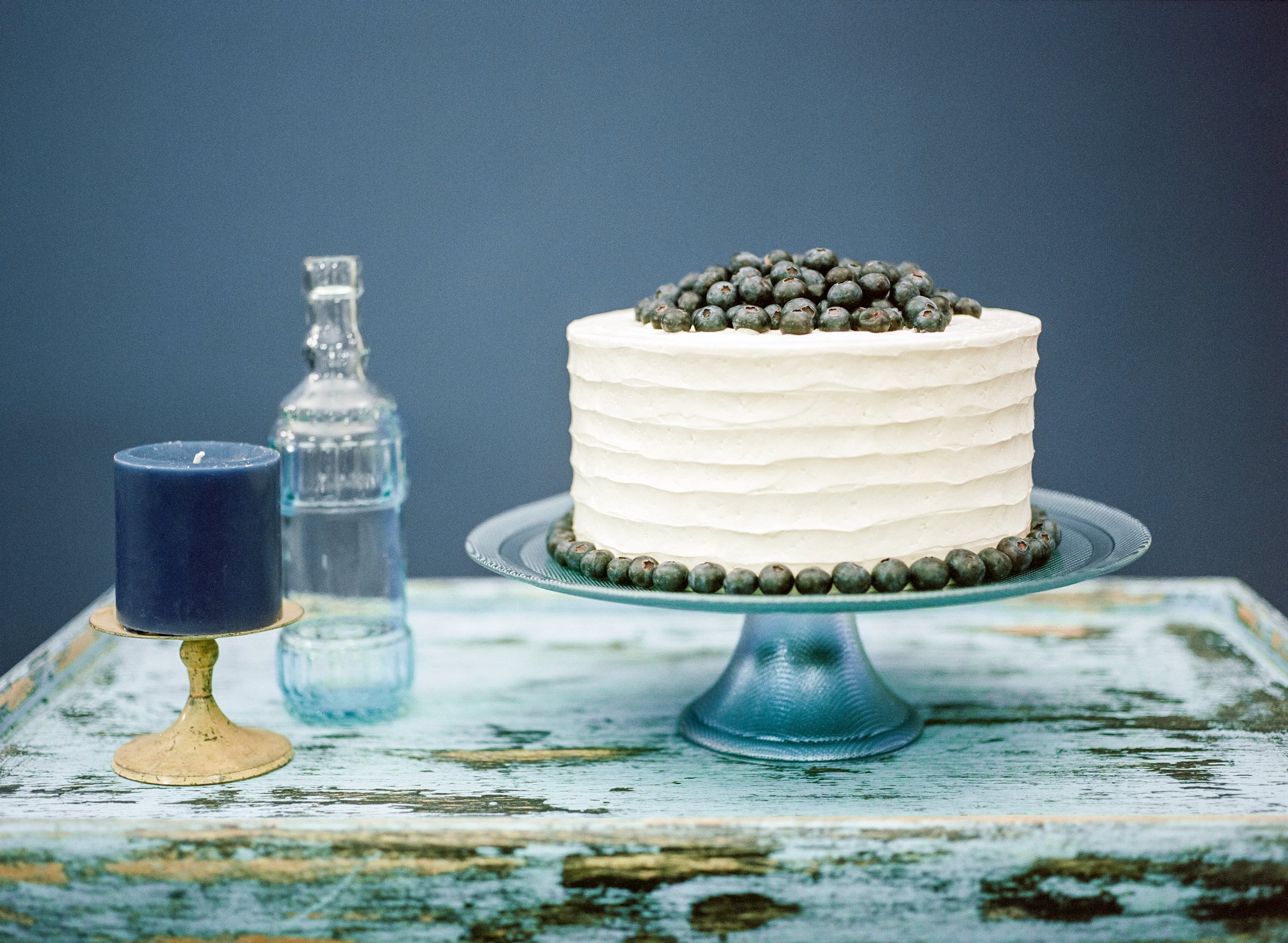 A white layered cake topped with blueberries on a blue dessert stand, surrounded by candles and glass bottles on a rustic wooden table.