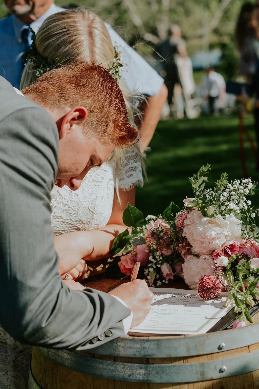 A couple signing a marriage license at an outdoor wedding ceremony, with a wedding bouquet of pink and white flowers resting on a wooden barrel.