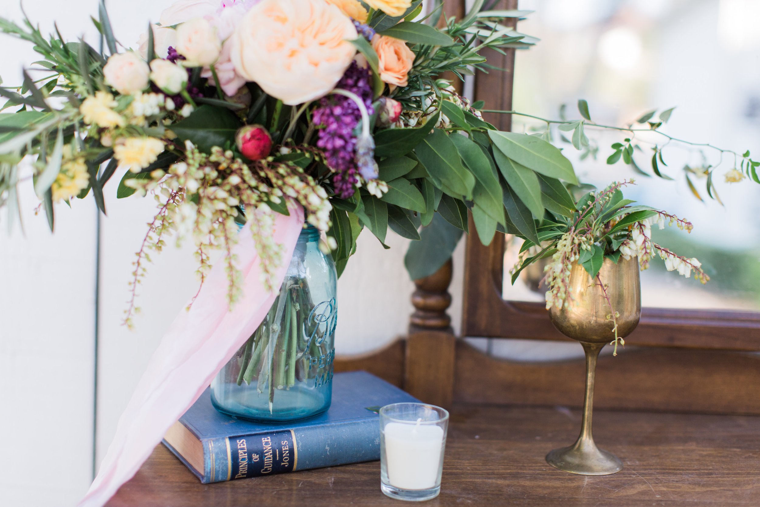 A floral arrangement in a blue glass Mason jar, sitting on a blue book, with pink and purple flowers and green leaves. A white candle in a glass holder and a brass vase with more green foliage and small flowers on a wooden table.