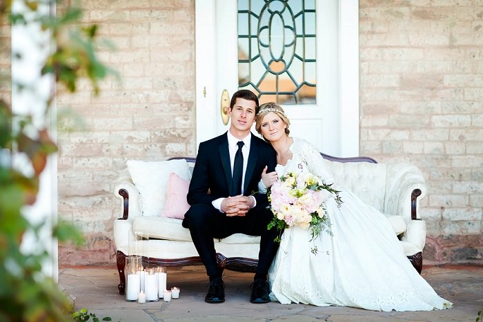 A bride and groom sitting on a vintage sofa in front of a brick wall, with candles on the ground nearby. The bride is holding a large bouquet of flowers.