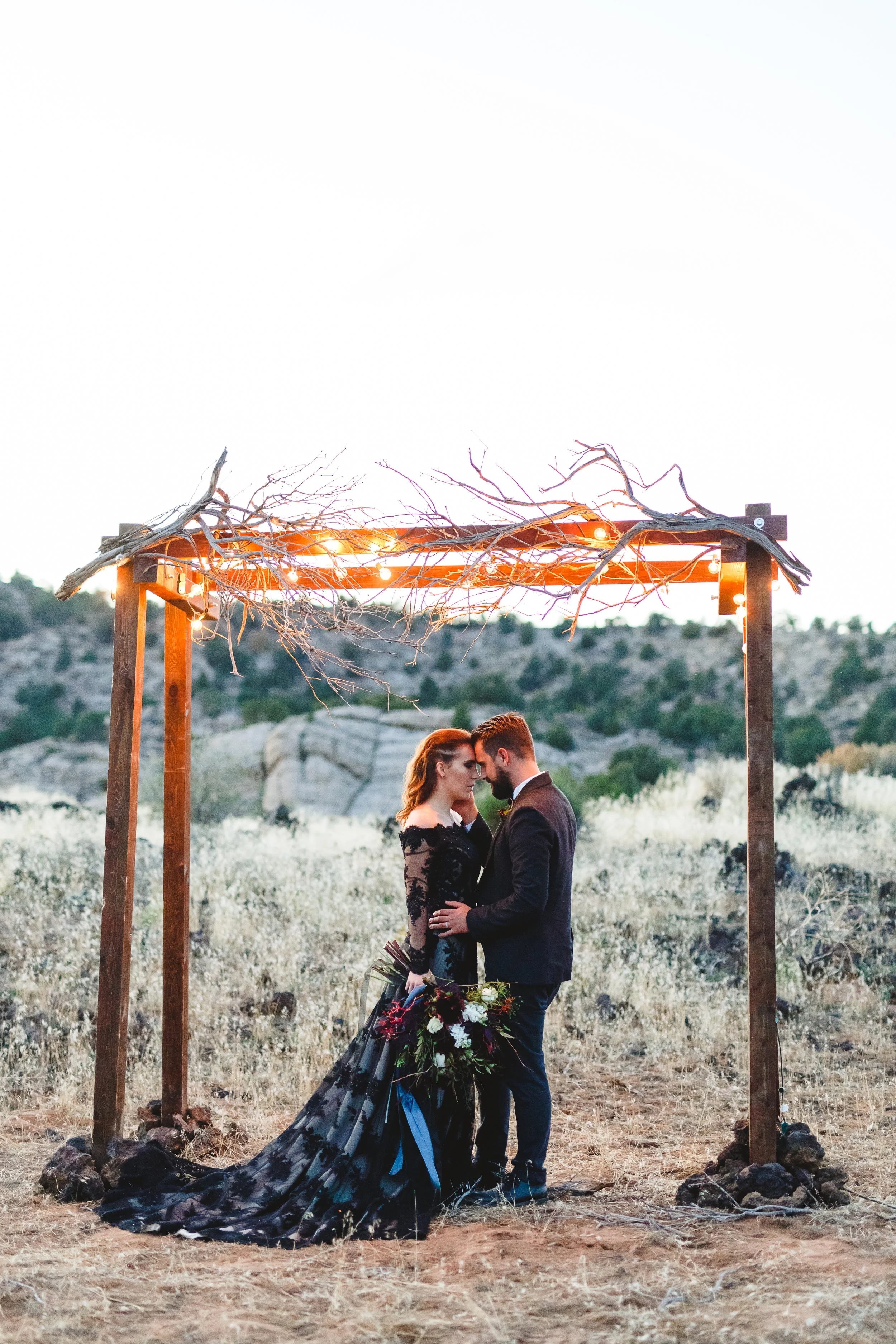 A romantic couple sharing a close moment during their wedding ceremony outdoors at sunset, under a simple wooden arch decorated with branches and string lights. The bride wears a dark gown with a long train and holds a bouquet, while the groom is dre