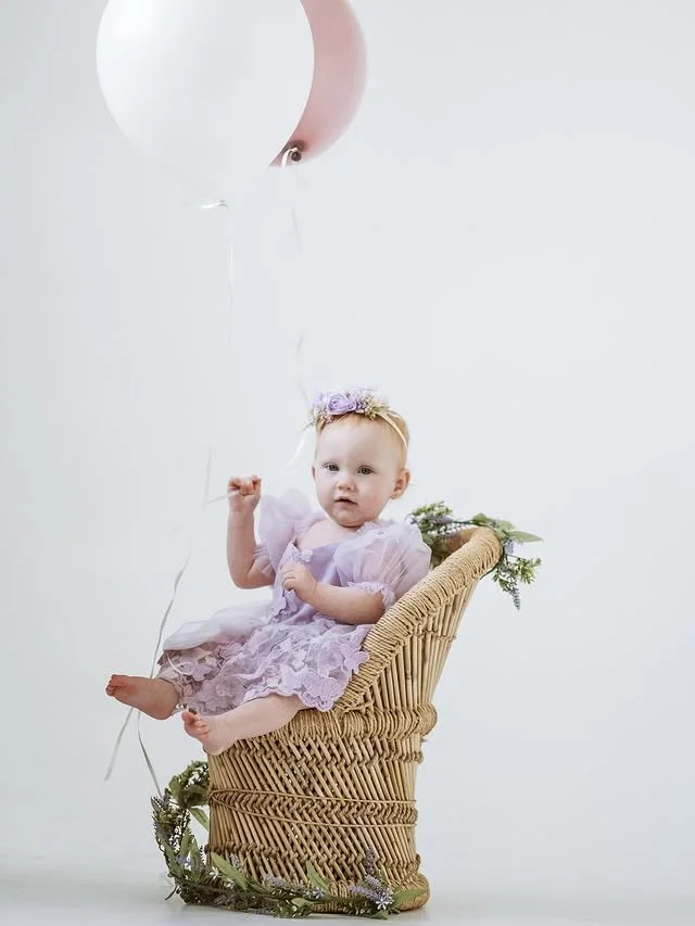 A young child in a purple dress sitting in a wicker basket with balloons above her, decorated with greenery.