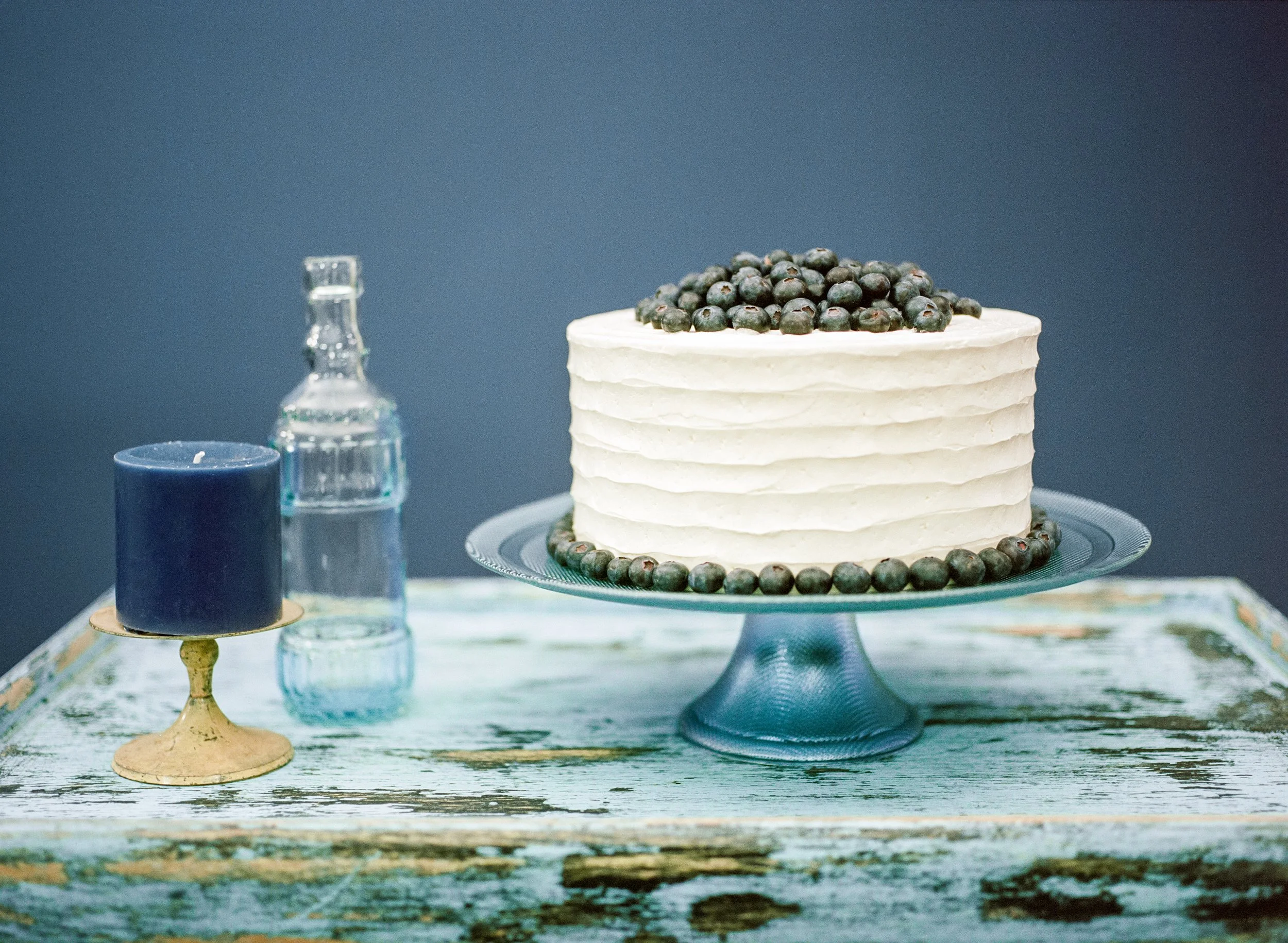 A tall, layered cake decorated with blueberries on top and around the base, displayed on a light blue cake stand. The background is a plain, muted blue, with a white and wooden table surface.
