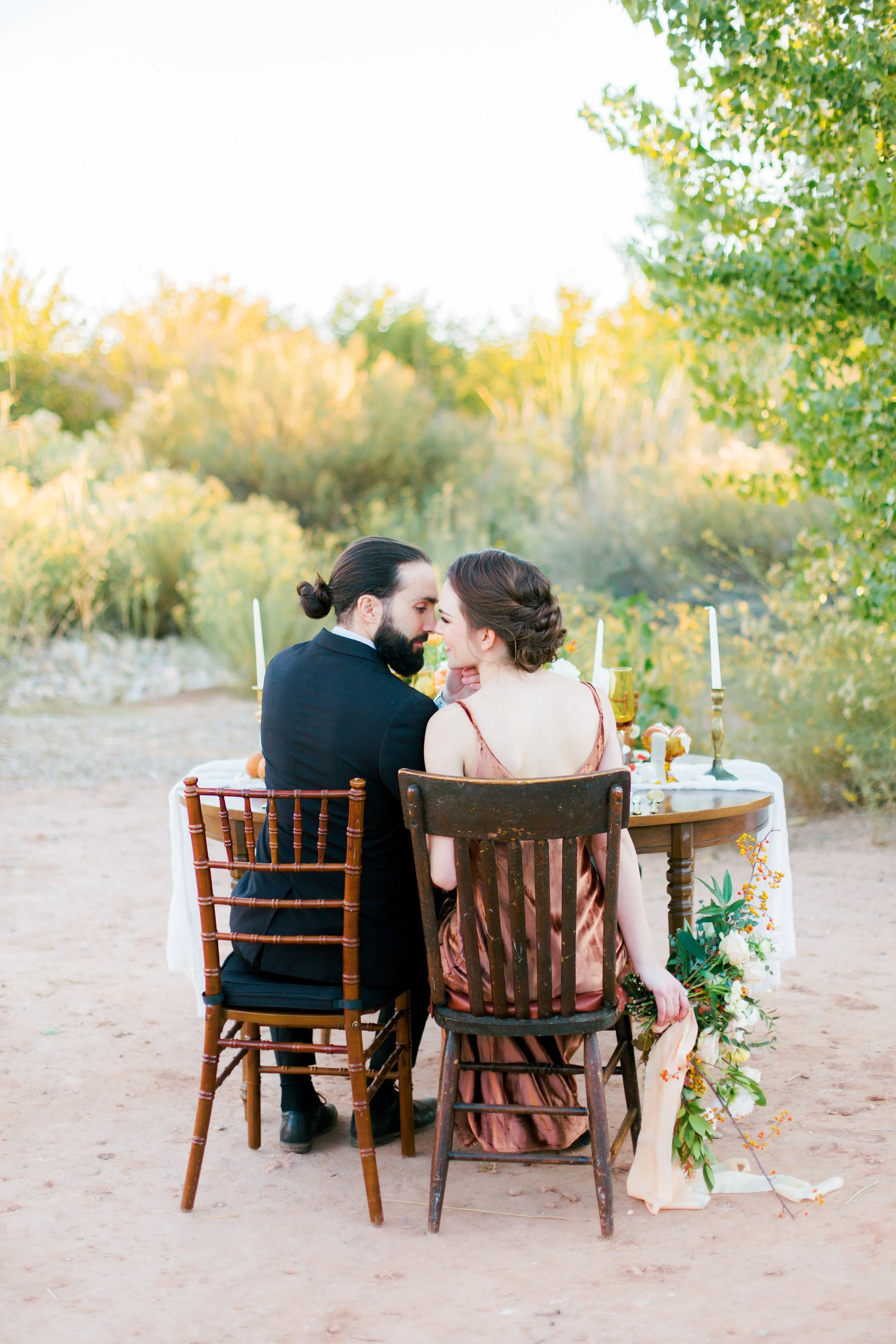 A couple sitting at a table outdoors, sharing a romantic moment with foreheads touching, surrounded by lush greenery and candlelit decor.