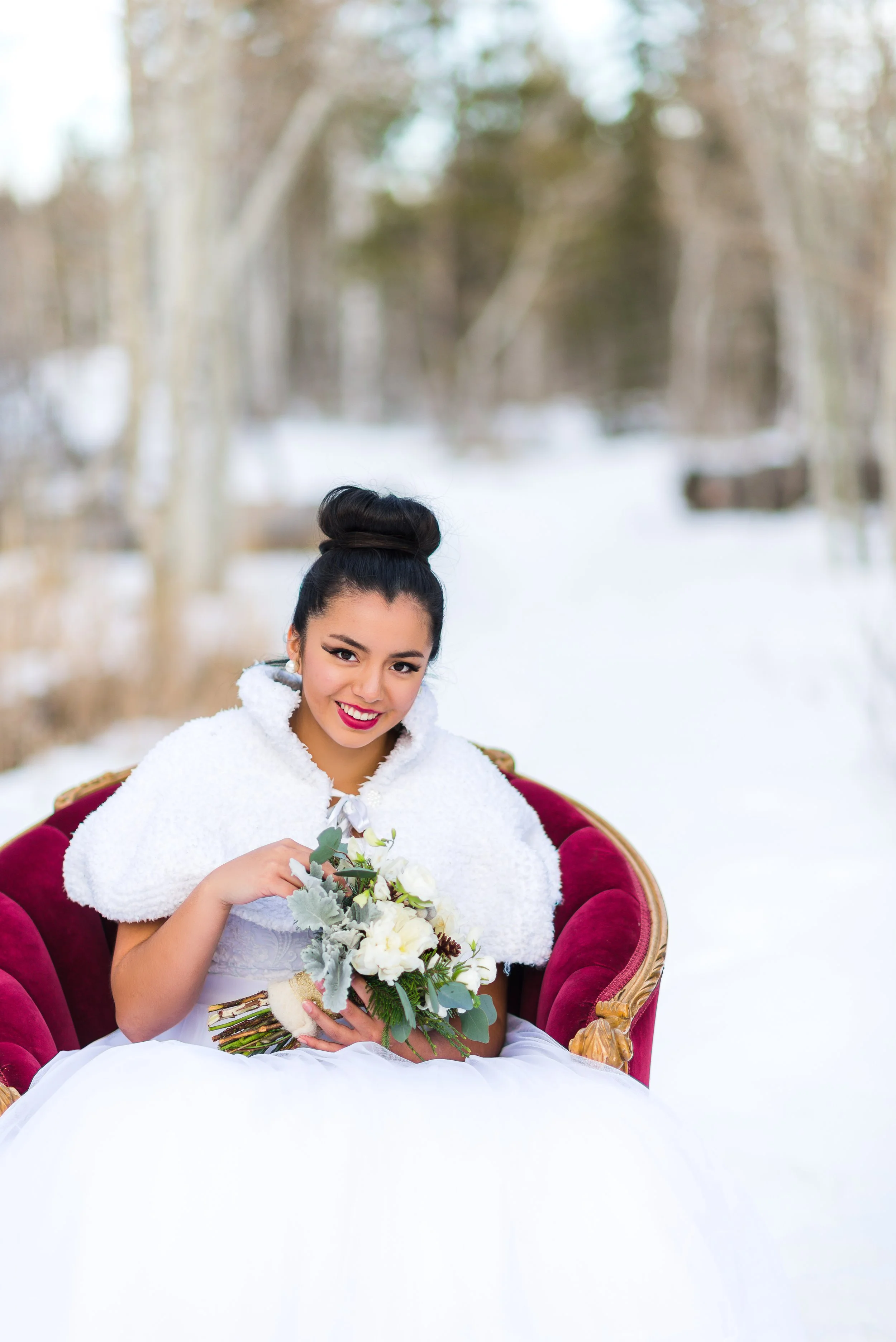 A woman dressed in a white outfit with a fluffy white cape, sitting on a red velvet chair outdoors in a snowy forest, holding a bouquet of white flowers and green foliage, smiling at the camera.