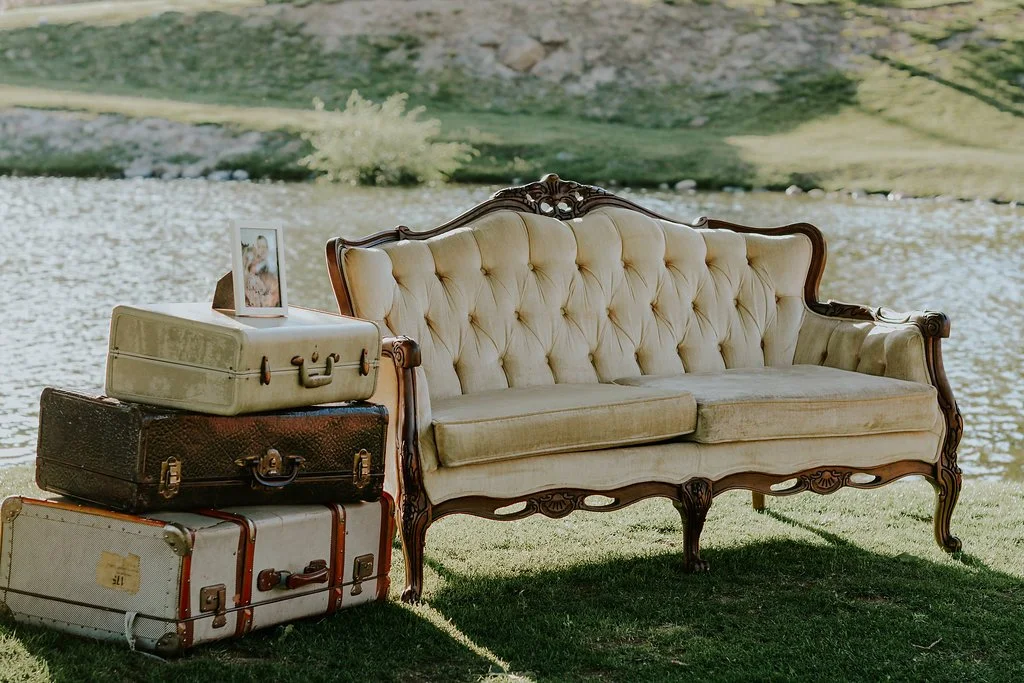 Vintage cream-colored sofa with wooden frame beside a stack of four suitcases near a lake.
